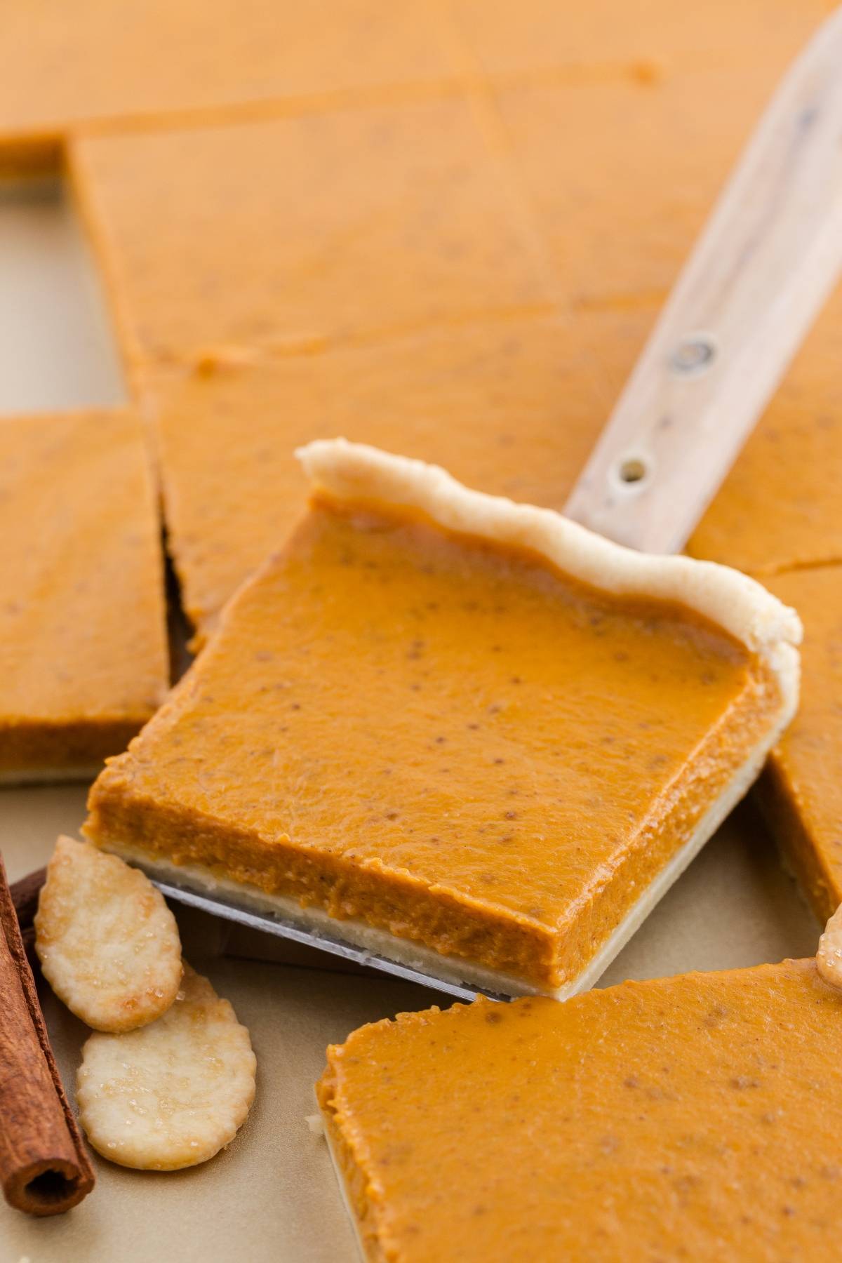 A slice of pumpkin pie bar on a spatula, surrounded by cinnamon sticks and pie crust cookies.