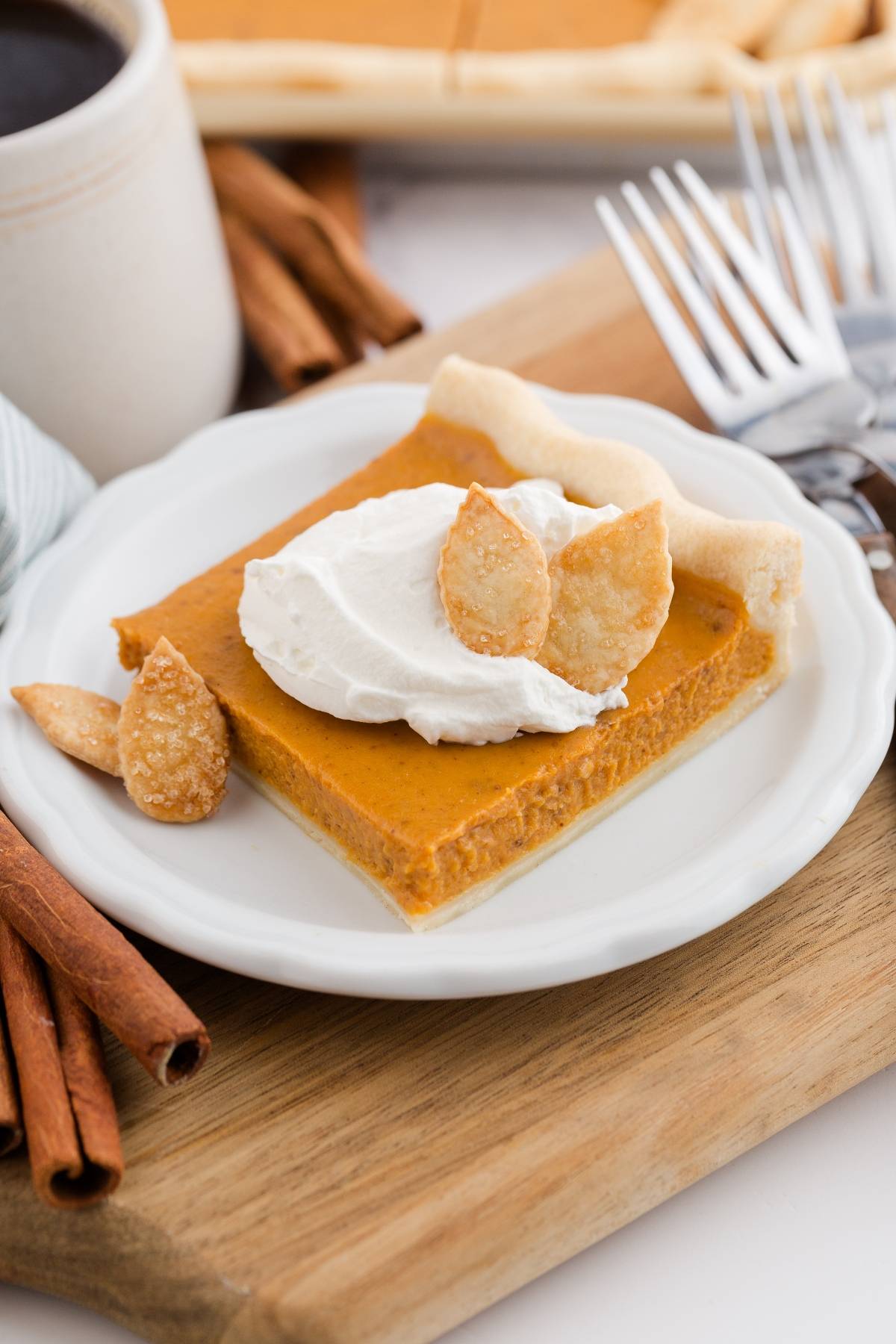A square pumpkin pie bar with whipped cream and pastry leaves on a white plate, next to forks and cinnamon sticks.