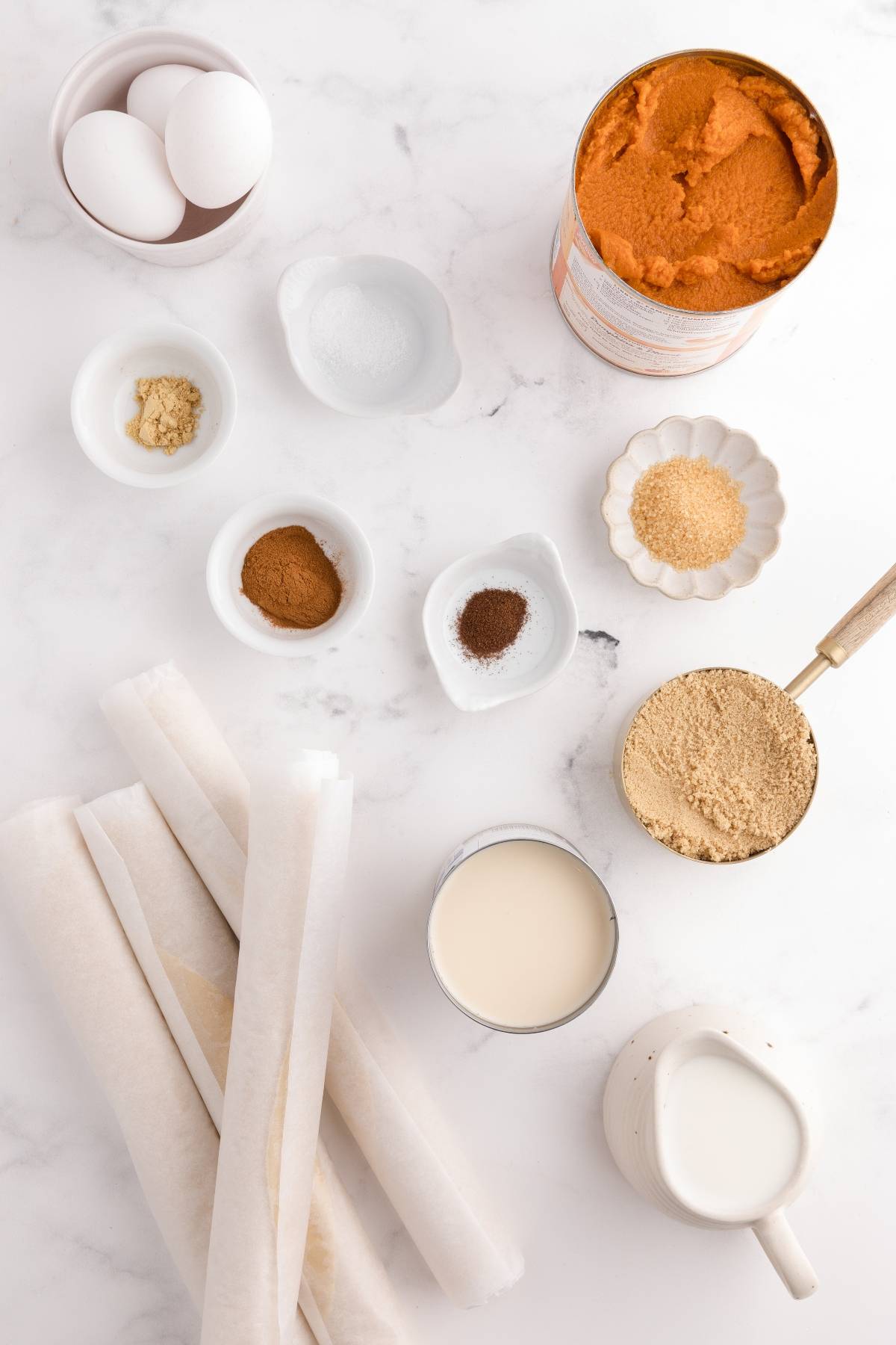 Baking ingredients on a white surface: eggs, pumpkin, spices, brown sugar, parchment paper, and milk.