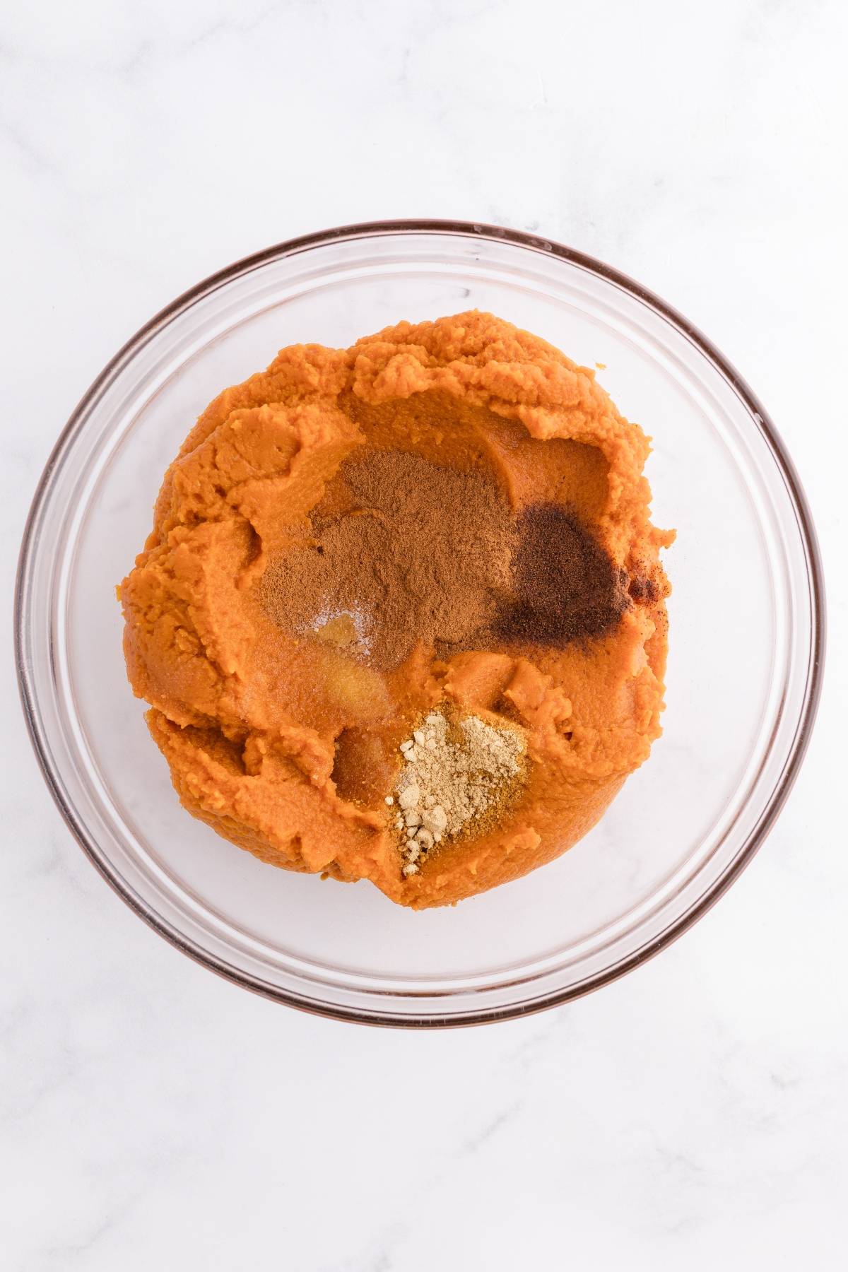 A glass bowl filled with pumpkin puree and various spices on a white surface.