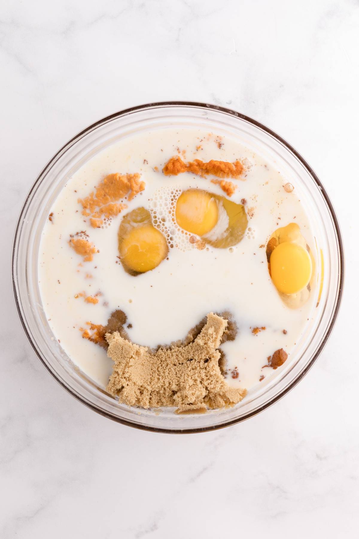 A glass bowl with eggs, milk, brown sugar, and spices on a white marble surface.