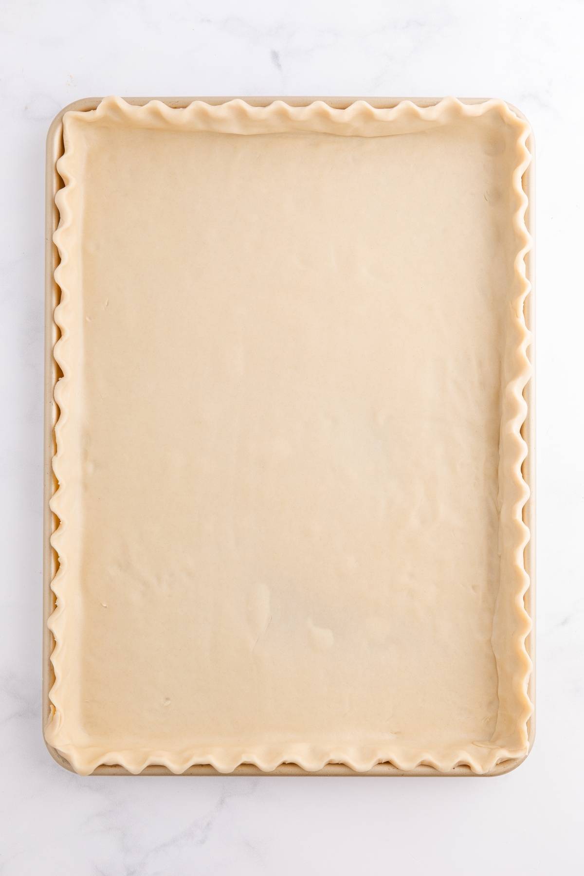 Unbaked rectangular pie crust with crimped edges in a baking sheet on a white surface.