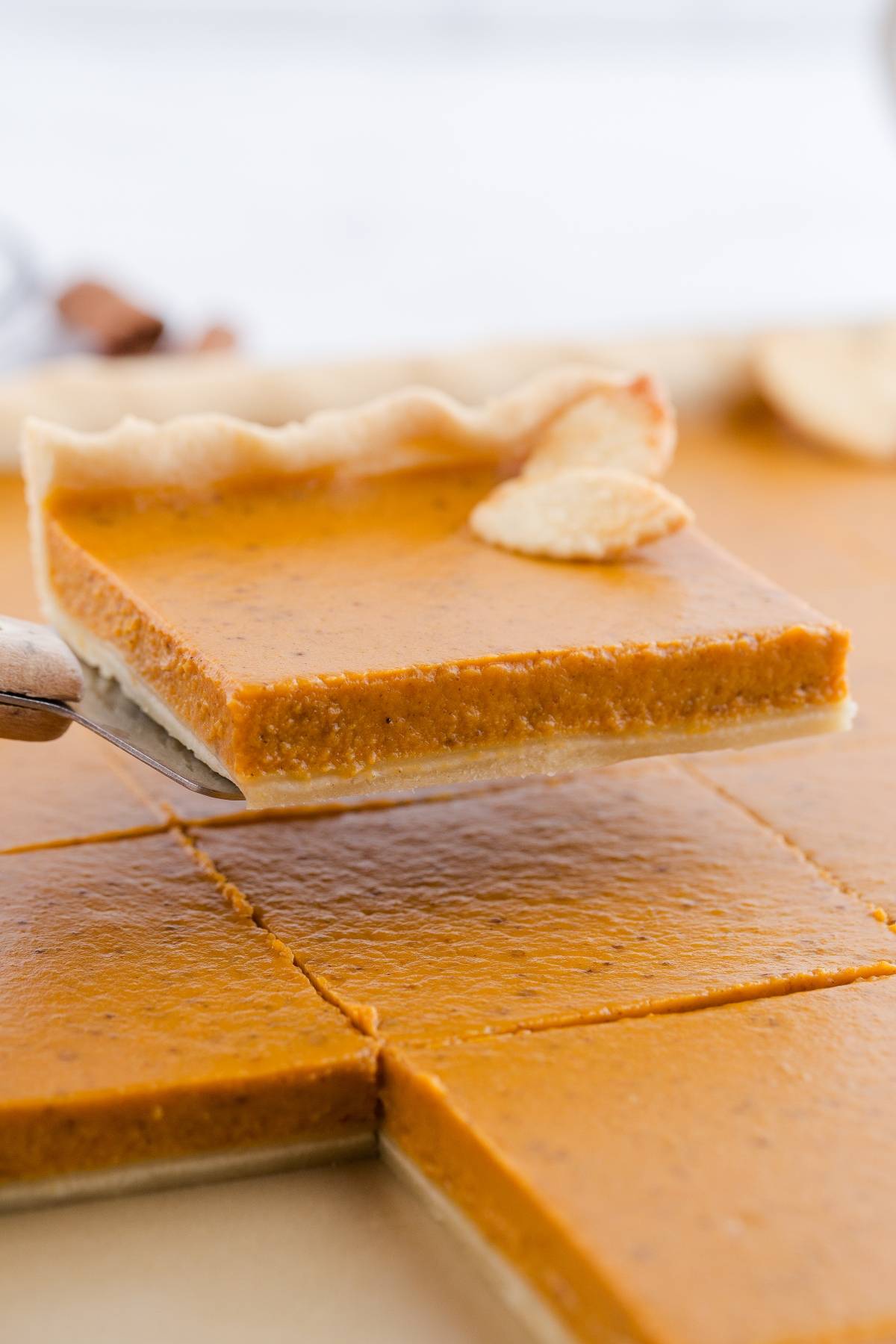 A square slice of pumpkin pie with a flaky crust is being lifted from a tray of neatly cut pieces.