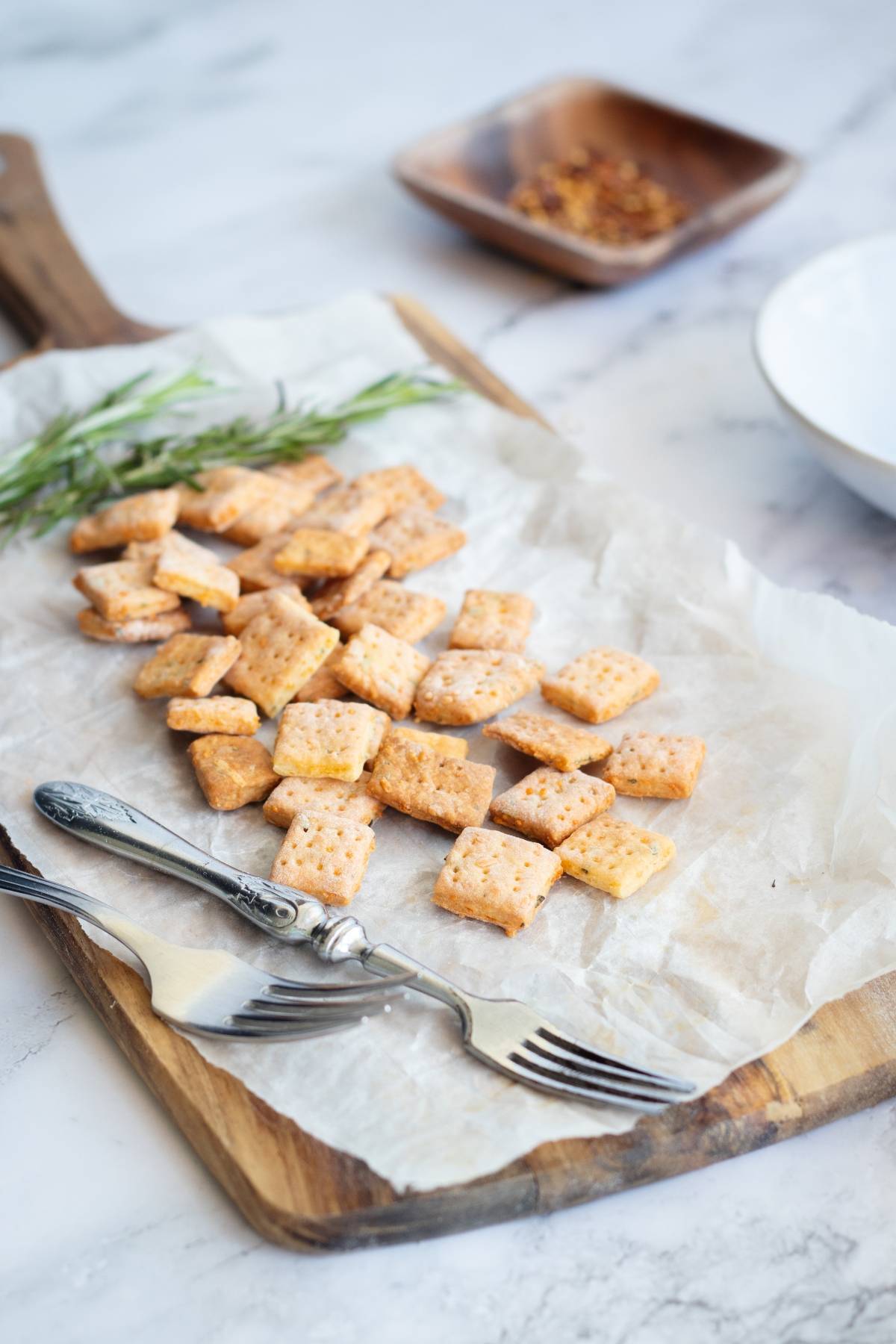 Square rosemary and cheddar crackers on parchment paper with two forks, fresh rosemary, and a bowl of spices atop a wooden board—an easy cracker recipe perfect for homemade snacking.