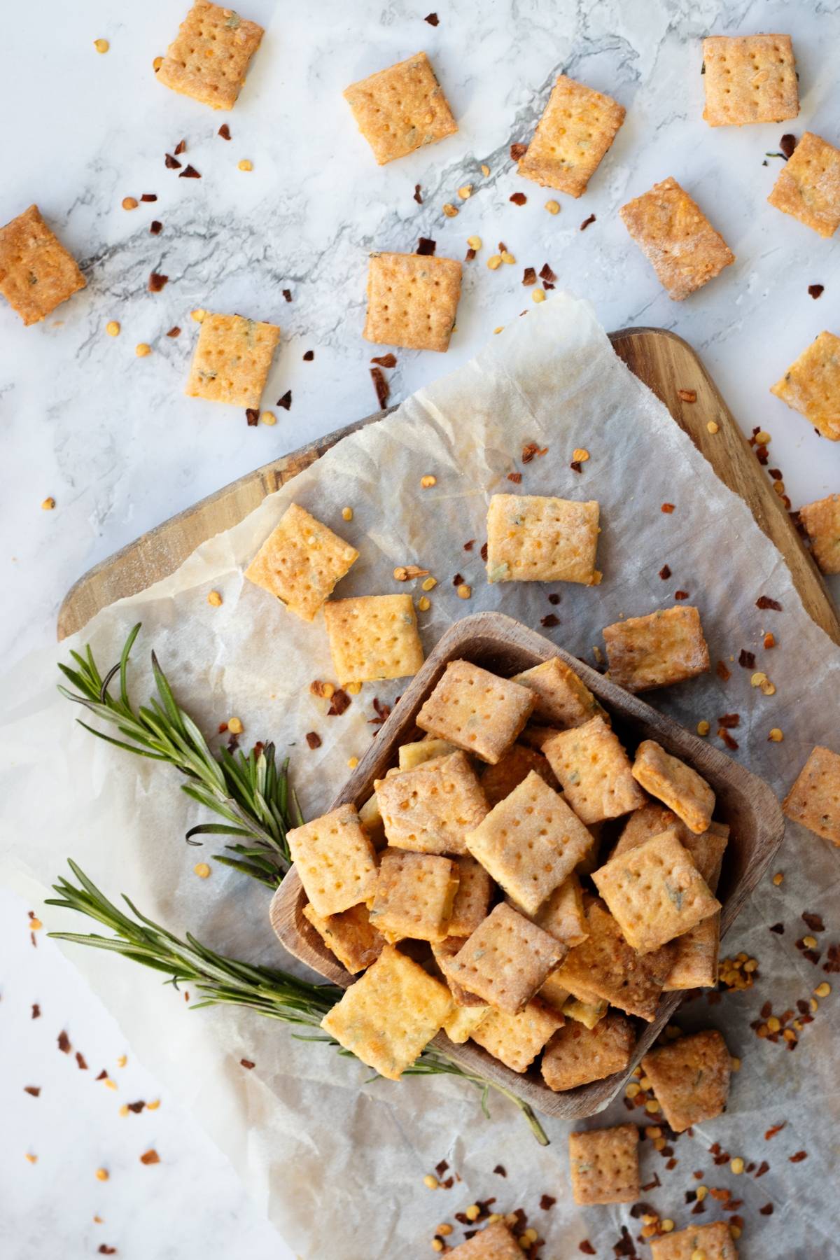 Square rosemary cheddar cheese crackers on parchment paper, garnished with rosemary, are scattered across a marble surface.