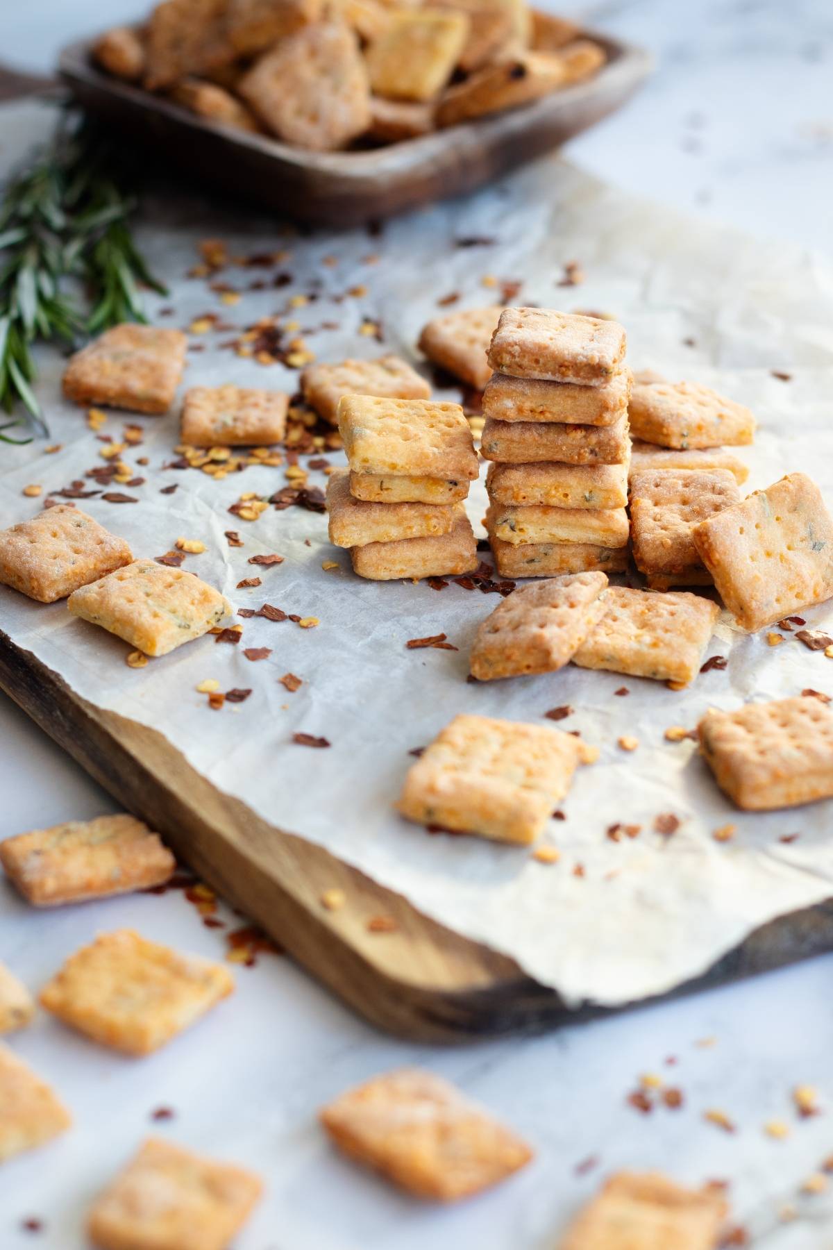 A stack of square rosemary cheddar cheese crackers on parchment paper, with more homemade cheese bites and herbs scattered around—an easy cracker recipe perfect for any snack spread.