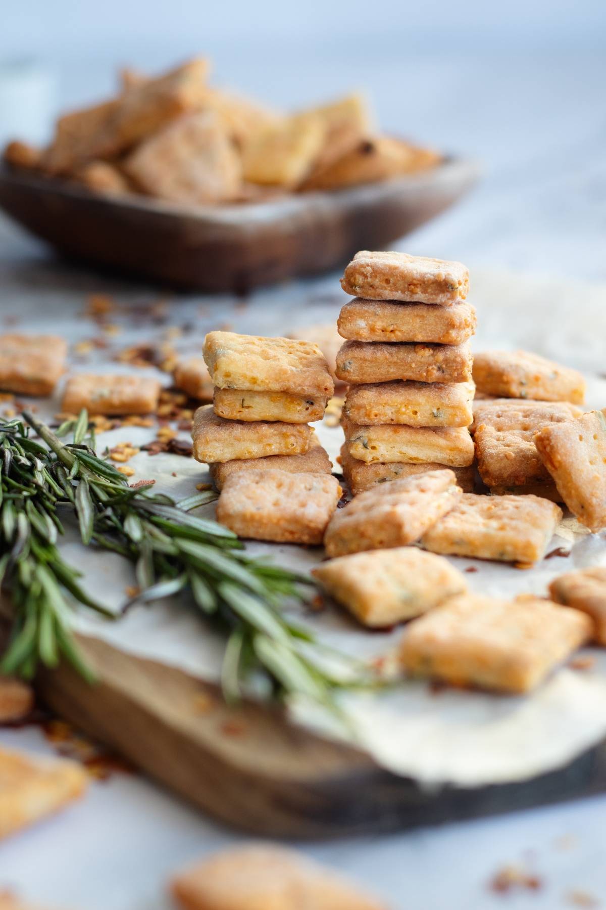 Stacked rosemary cheddar cheese crackers on a wooden board with fresh rosemary and more crackers in the background.
