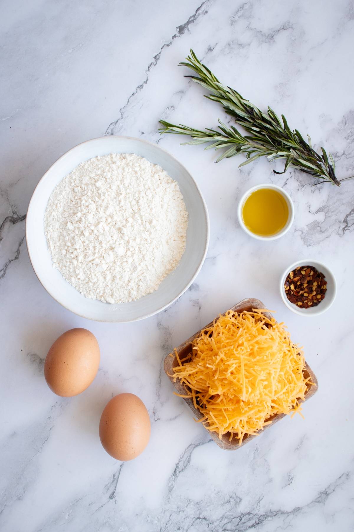 Bowl of flour, shredded cheese, two eggs, rosemary, olive oil, and spices on a marble surface—perfect ingredients for homemade rosemary cheddar cheese crackers.