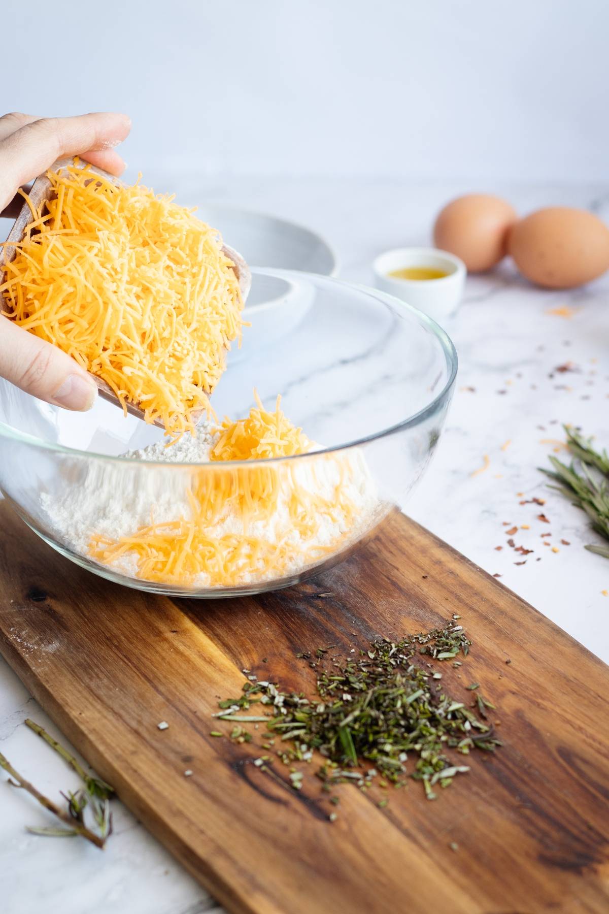 A hand adds shredded cheese to a glass bowl on a table with eggs, herbs, and oil nearby—perfect for making an easy cracker recipe or delicious homemade cheese bites.