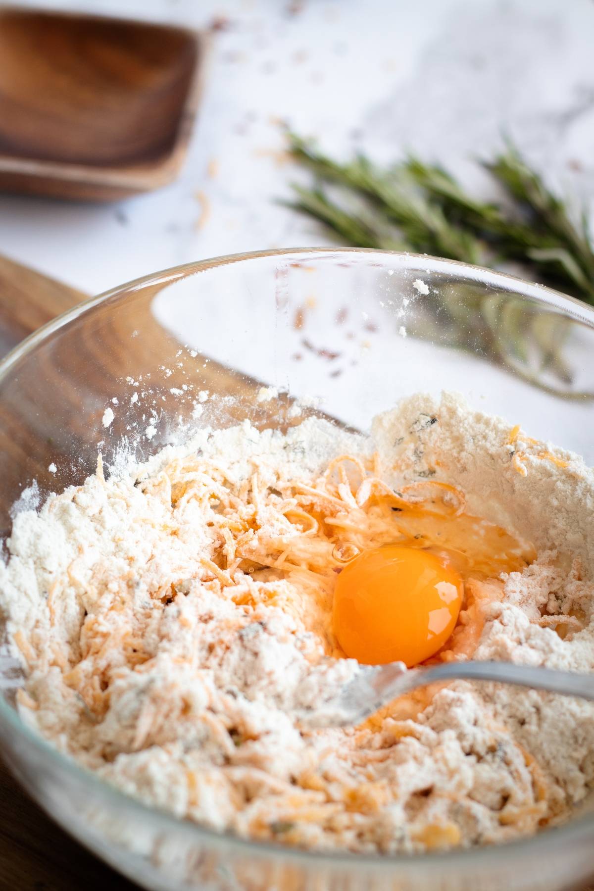 A glass bowl with flour, shredded cheese, and a cracked egg, ready to be mixed for easy cracker recipe prep on the kitchen counter.