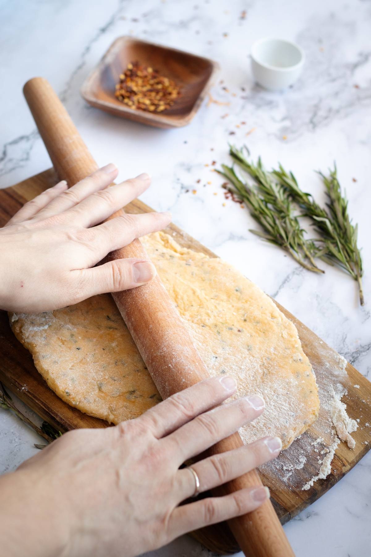 Hands rolling out herbed dough for easy cracker recipe with a rolling pin on a wooden board, rosemary and spices nearby.