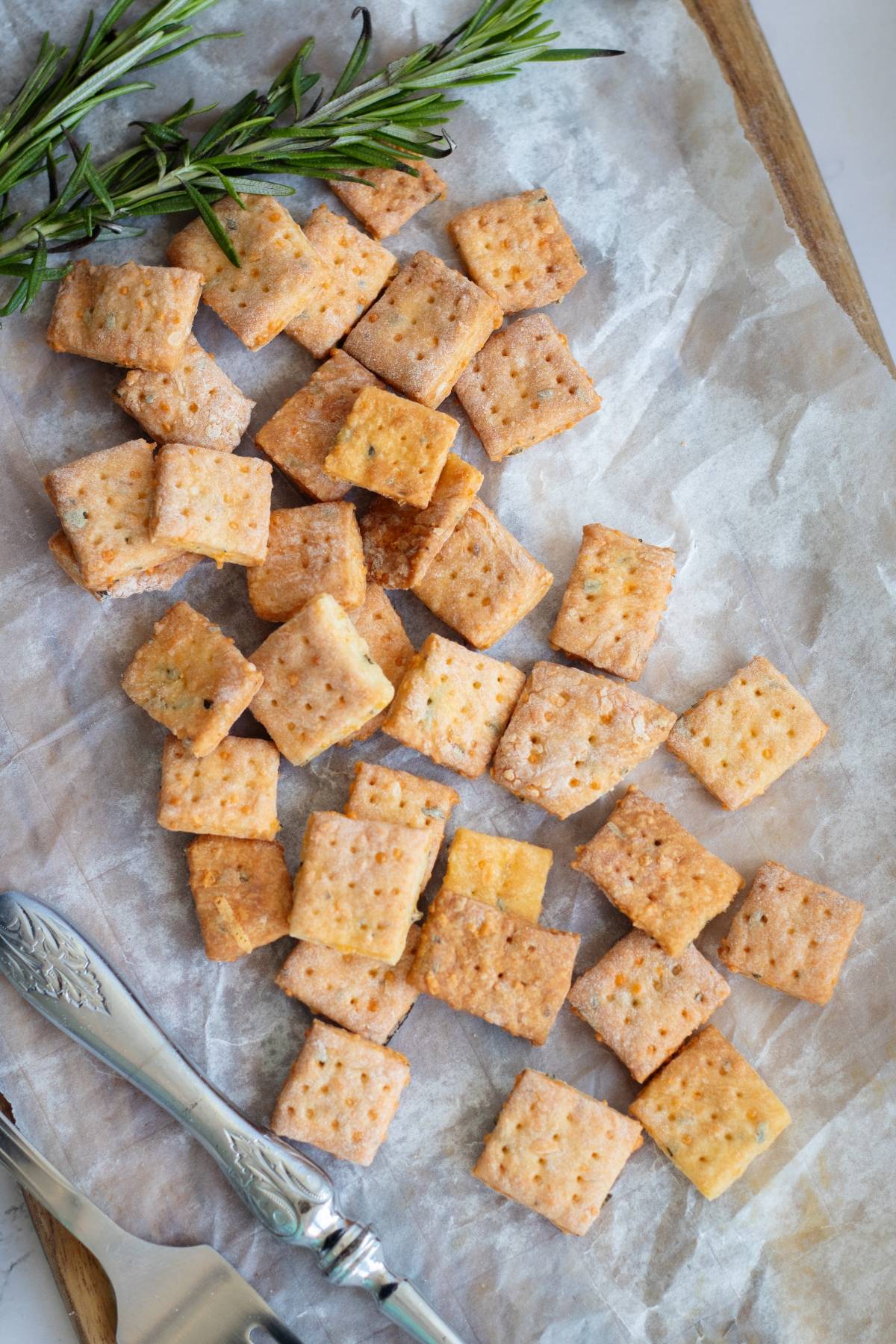 Rosemary and cheddar crackers and a sprig of rosemary rest on parchment paper, with a silver knife and fork nearby.