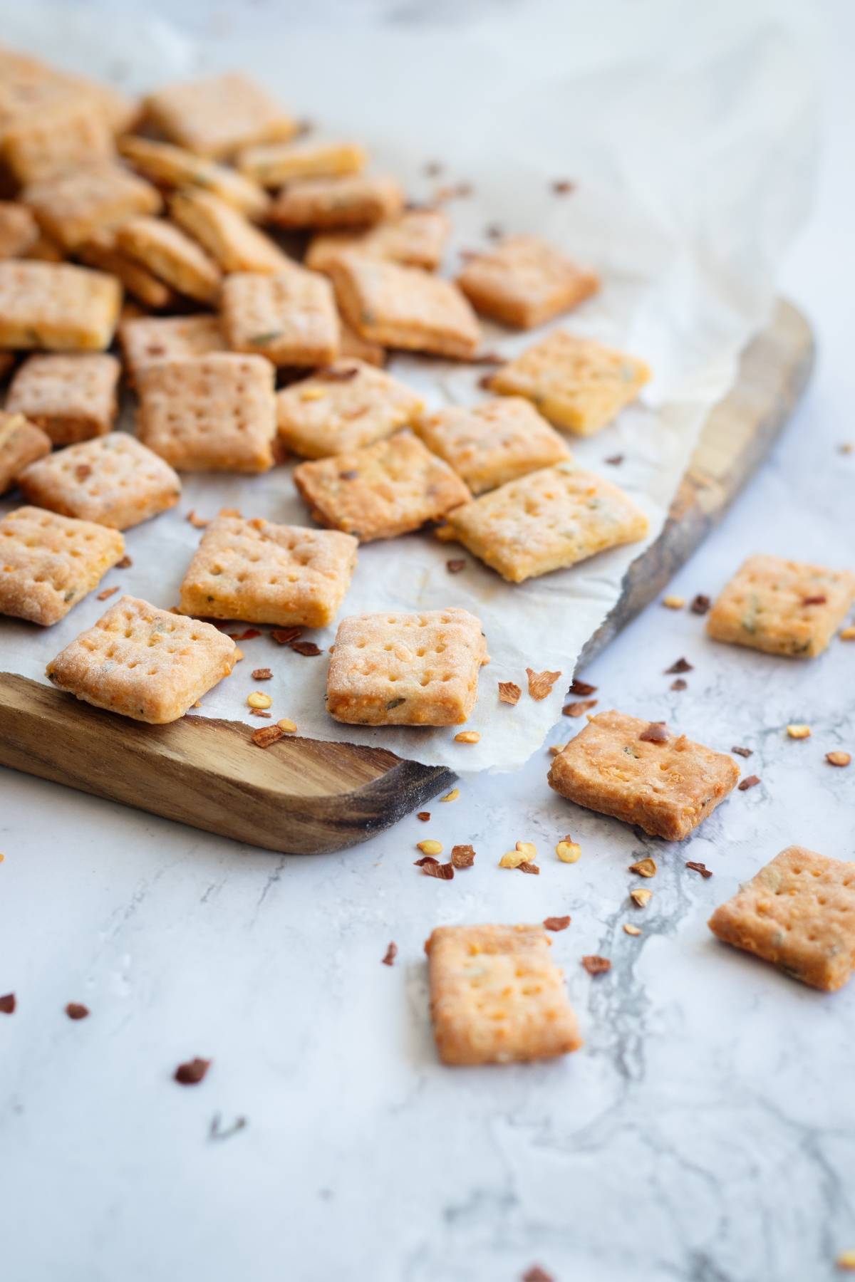 Square rosemary and cheddar crackers are scattered on parchment paper atop a wooden board, with some crumbs on a marble surface—a perfect showcase for this homemade cracker recipe.