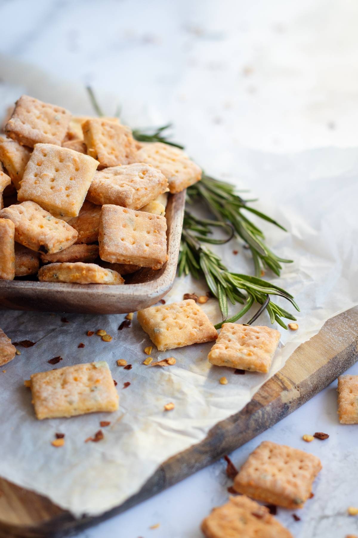 Rosemary and cheddar crackers on parchment paper, sprinkled with herbs and chili flakes—some in a wooden bowl, others scattered around. An easy cracker recipe perfect for snacking or sharing.