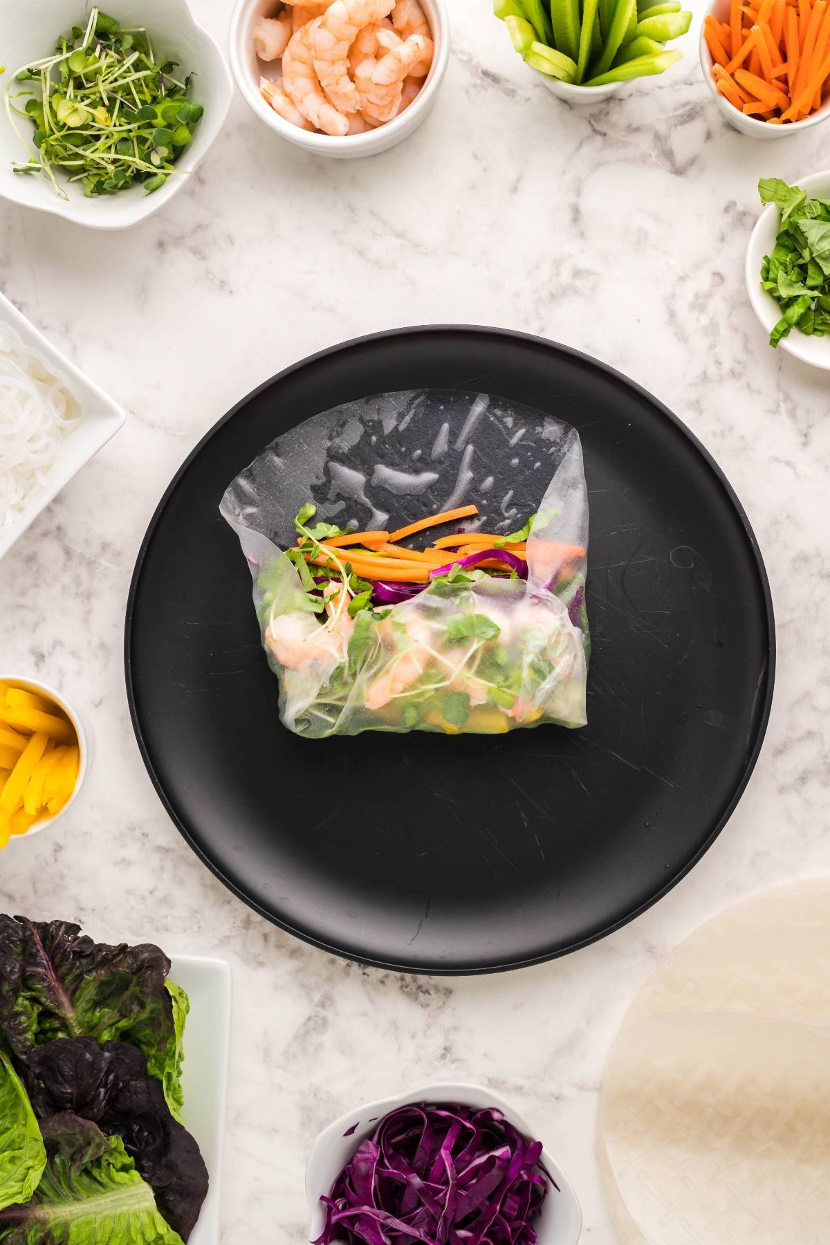 A fresh Vietnamese spring roll with colorful veggies sits on a black plate, surrounded by bowls of ingredients and a tangy dipping sauce for spring rolls.