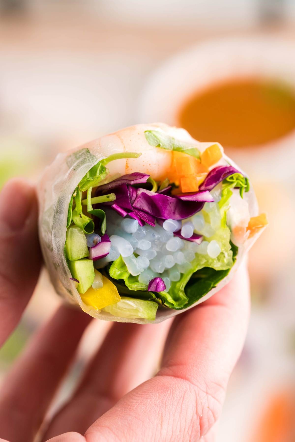A hand holds a fresh shrimp spring roll filled with colorful vegetables and clear noodles, perfect for dipping in spicy garlic sauce.