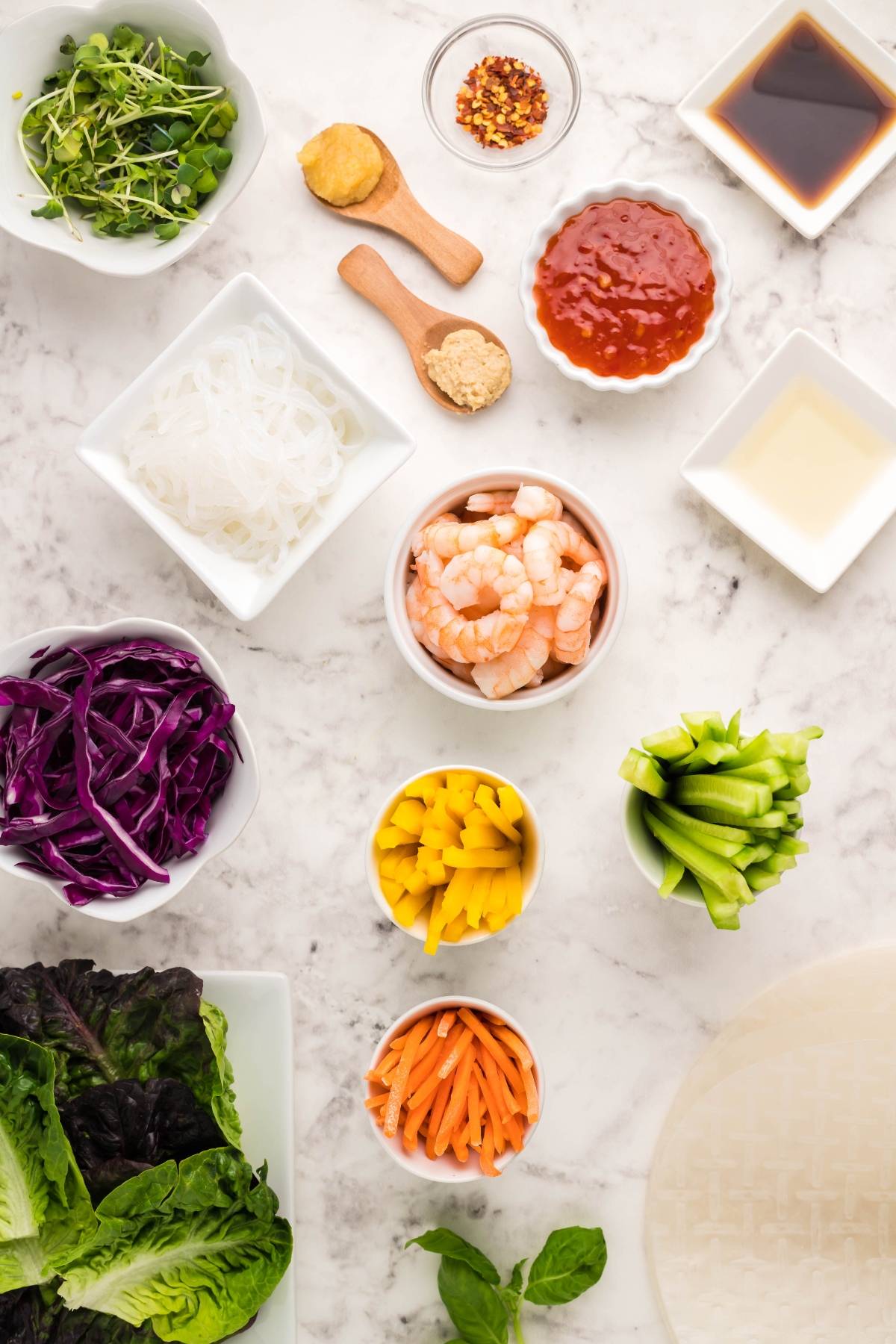 A variety of ingredients for a shrimp spring roll recipe—shrimp, vegetables, noodles, and dipping sauce for spring rolls—are artfully arranged on a marble surface.