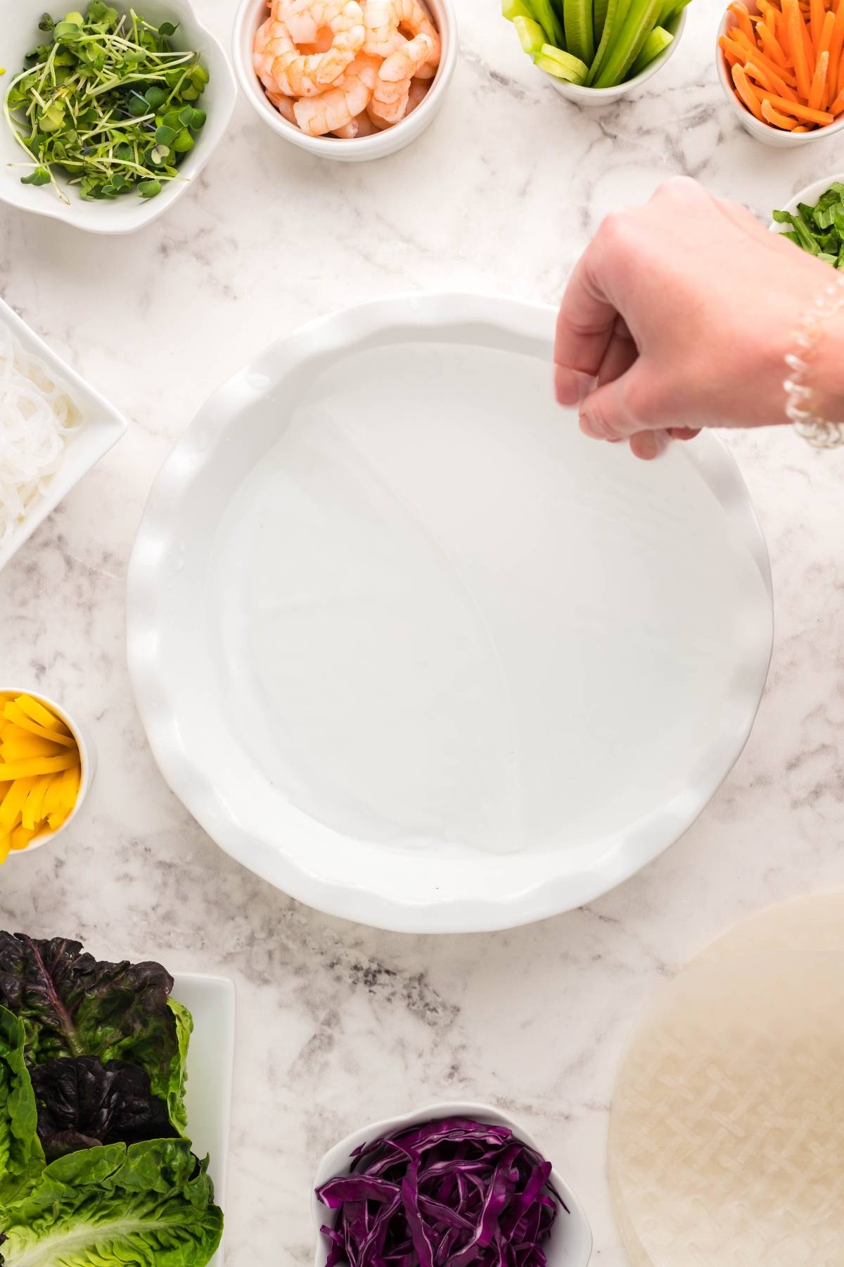 A hand dips a rice paper wrapper into water, surrounded by fresh vegetables and shrimp—classic ingredients for fresh Vietnamese spring rolls.