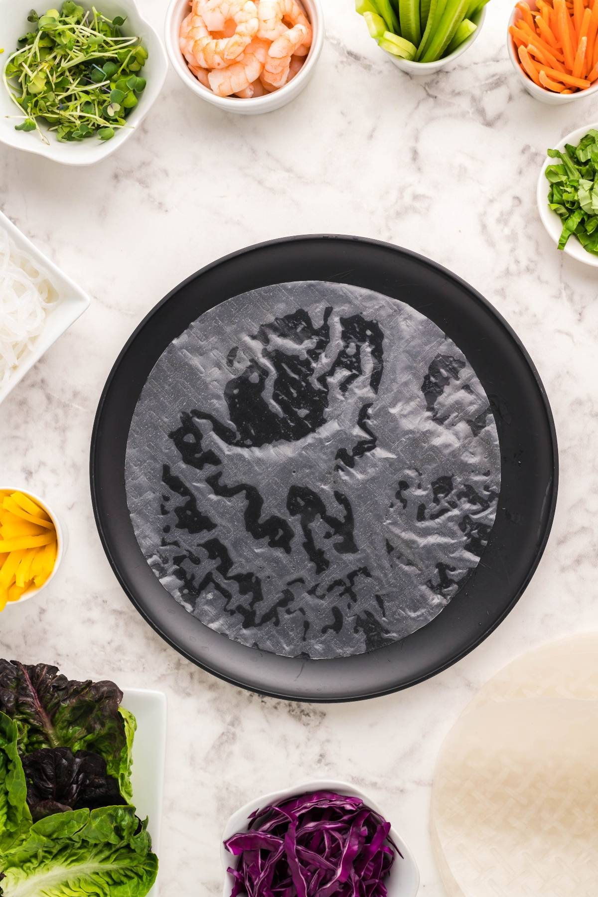 A rice paper sheet on a black plate is surrounded by bowls of shrimp, vegetables, and herbs—perfect for assembling your favorite shrimp spring rolls and pairing with a delicious dipping sauce for spring rolls.