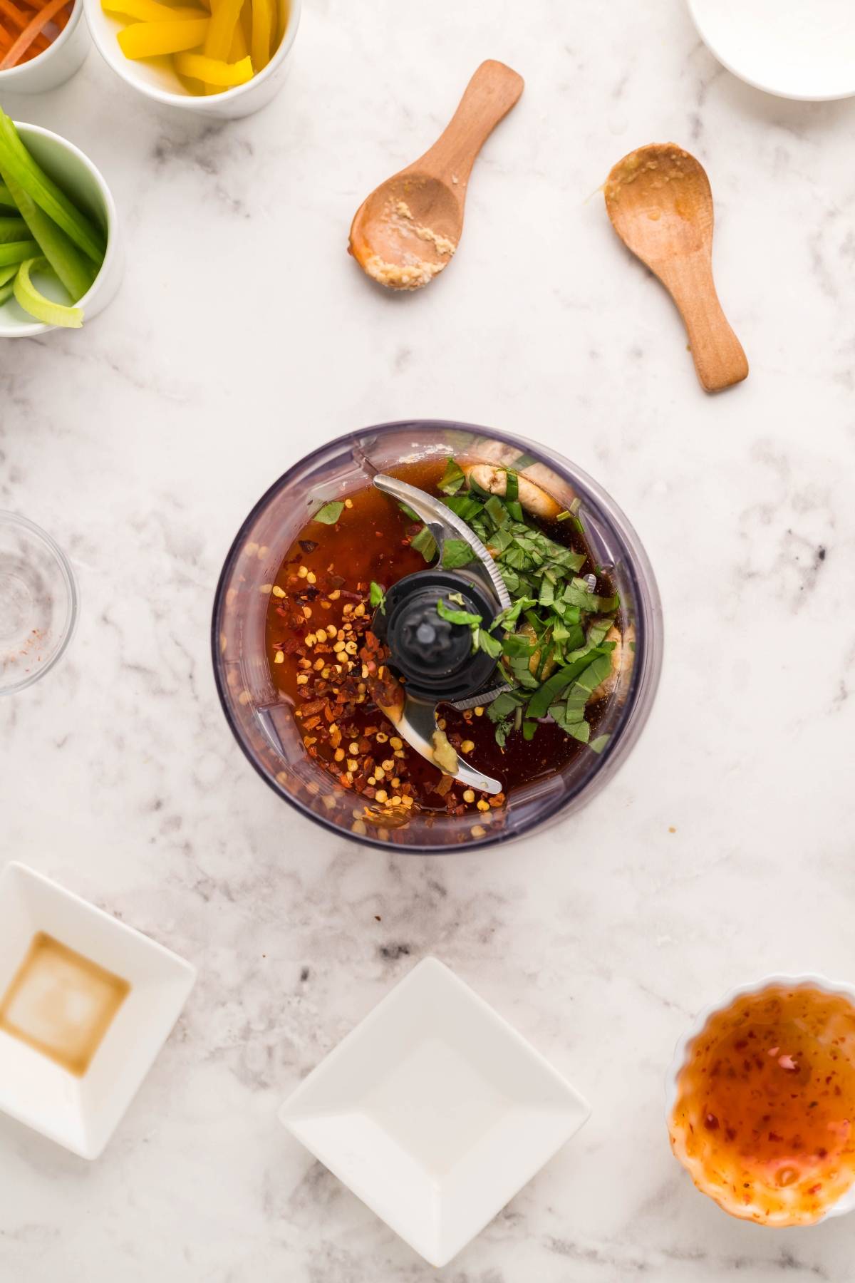 Top view of a food processor preparing dipping sauce for spring rolls, surrounded by fresh herbs and spices on a marble counter with bowls and wooden spoons nearby—perfect for your favorite fresh Vietnamese spring rolls.