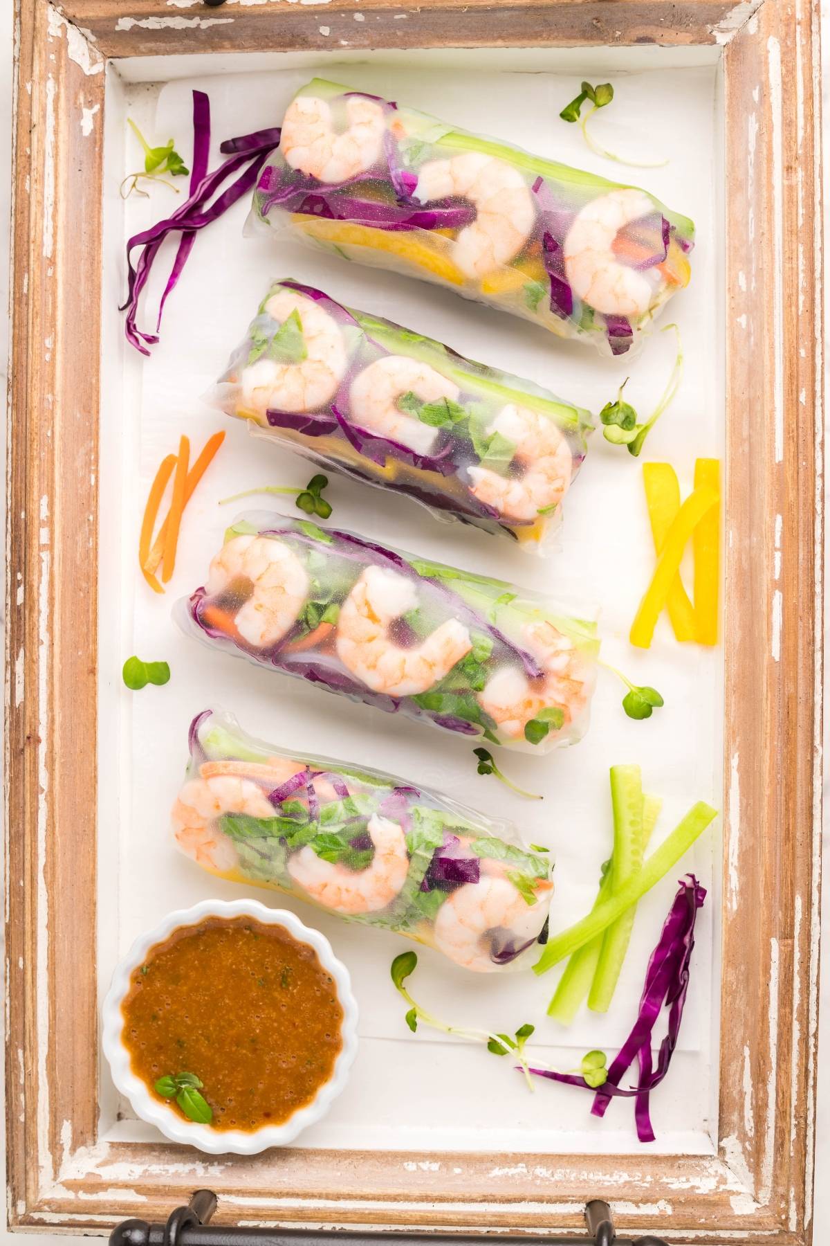 Four fresh Vietnamese spring rolls with colorful vegetables and shrimp on a tray, served with a spicy garlic sauce for dipping and veggie garnishes.