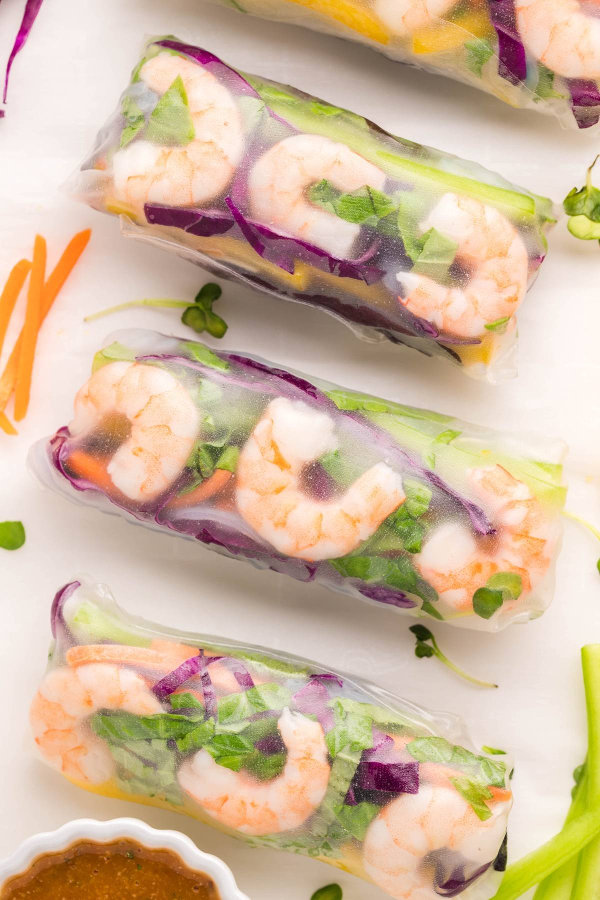 Three shrimp spring rolls with vegetables rest on a white surface, accompanied by a flavorful dipping sauce for spring rolls and shredded veggies nearby.
