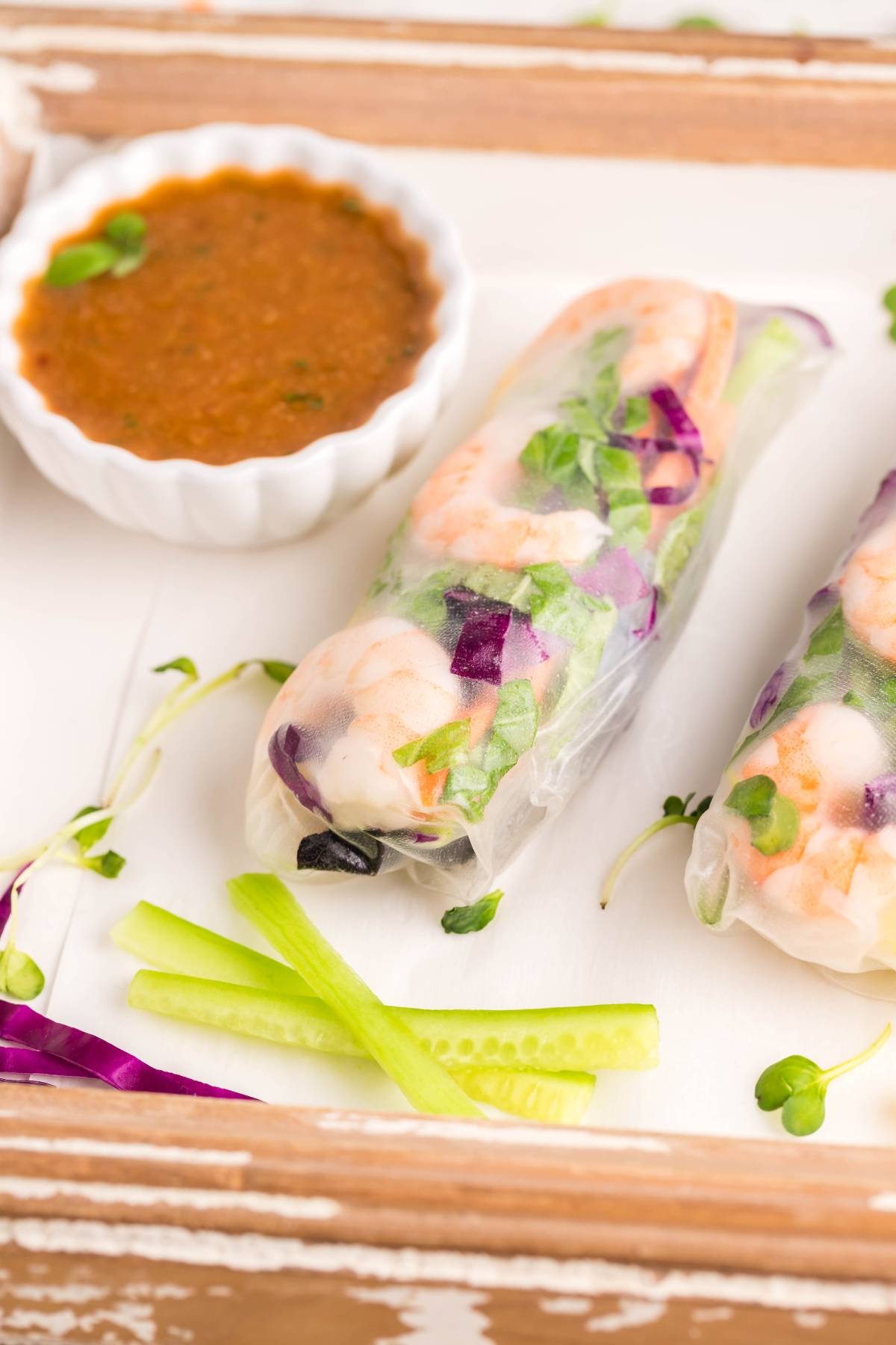 Two fresh Vietnamese spring rolls with shrimp and vegetables are served beside a small bowl of dipping sauce for spring rolls and cucumber slices.