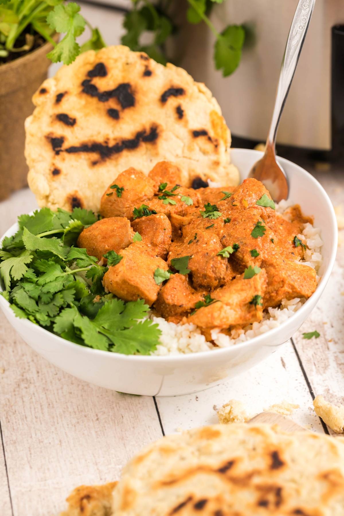 A bowl of slow cooker butter chicken on rice with fresh cilantro and naan bread, garnished with chopped herbs.