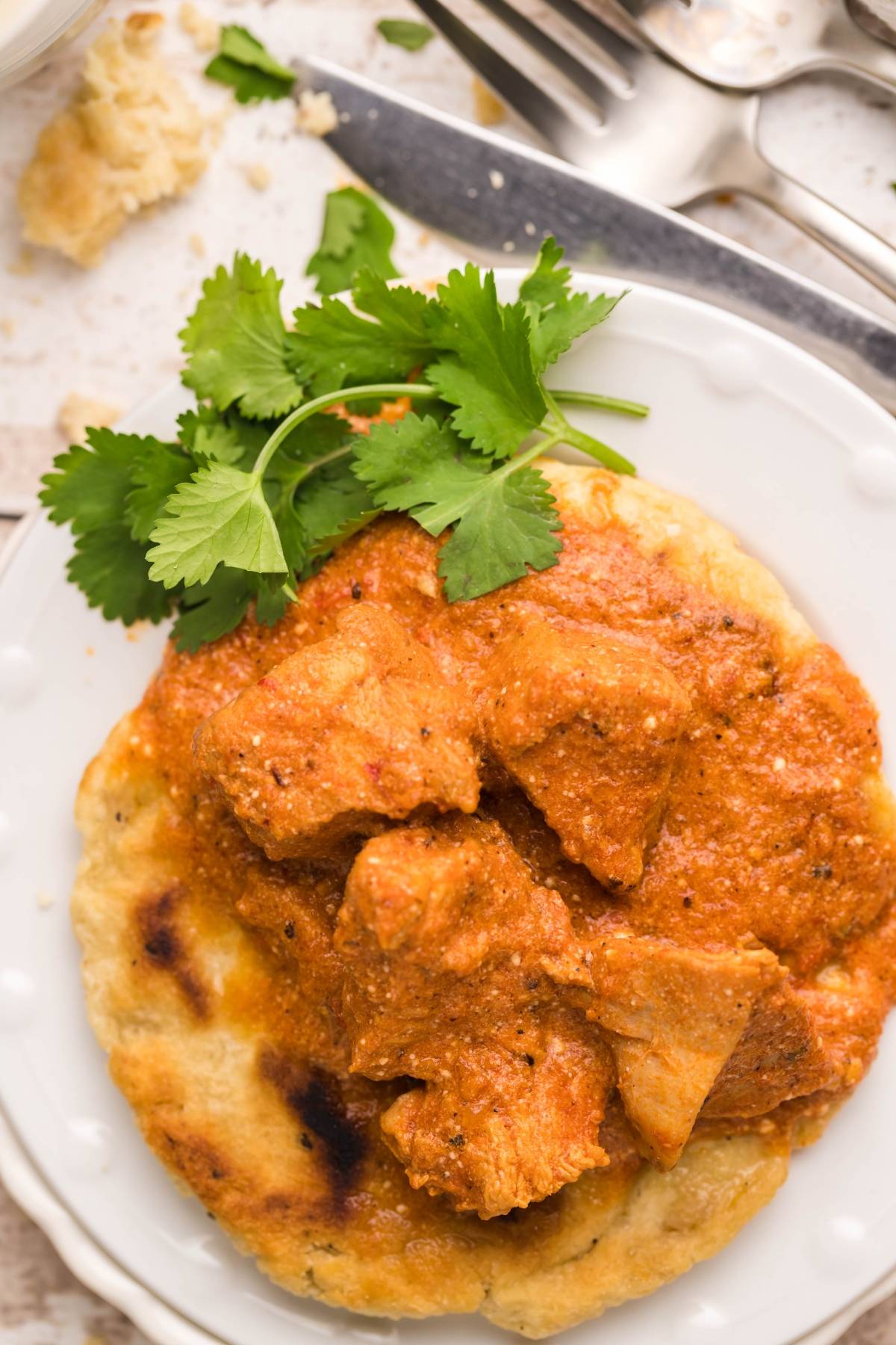 Tender slow cooker butter chicken on naan bread, garnished with fresh cilantro and served on a white plate with cutlery nearby.