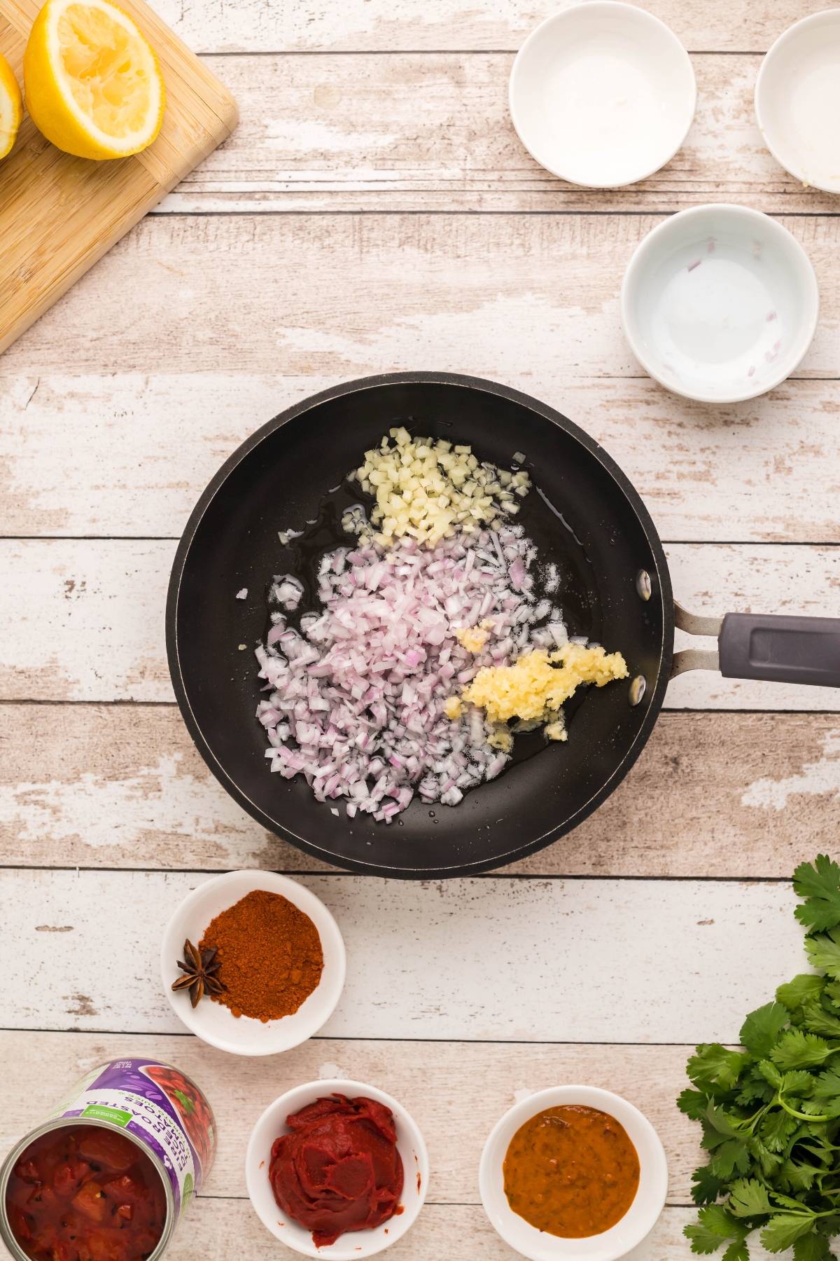 Chopped onions, garlic, and ginger in a frying pan surrounded by various spices and ingredients—a flavorful start for an Easy Slow Cooker Dairy Free Butter Chicken recipe.