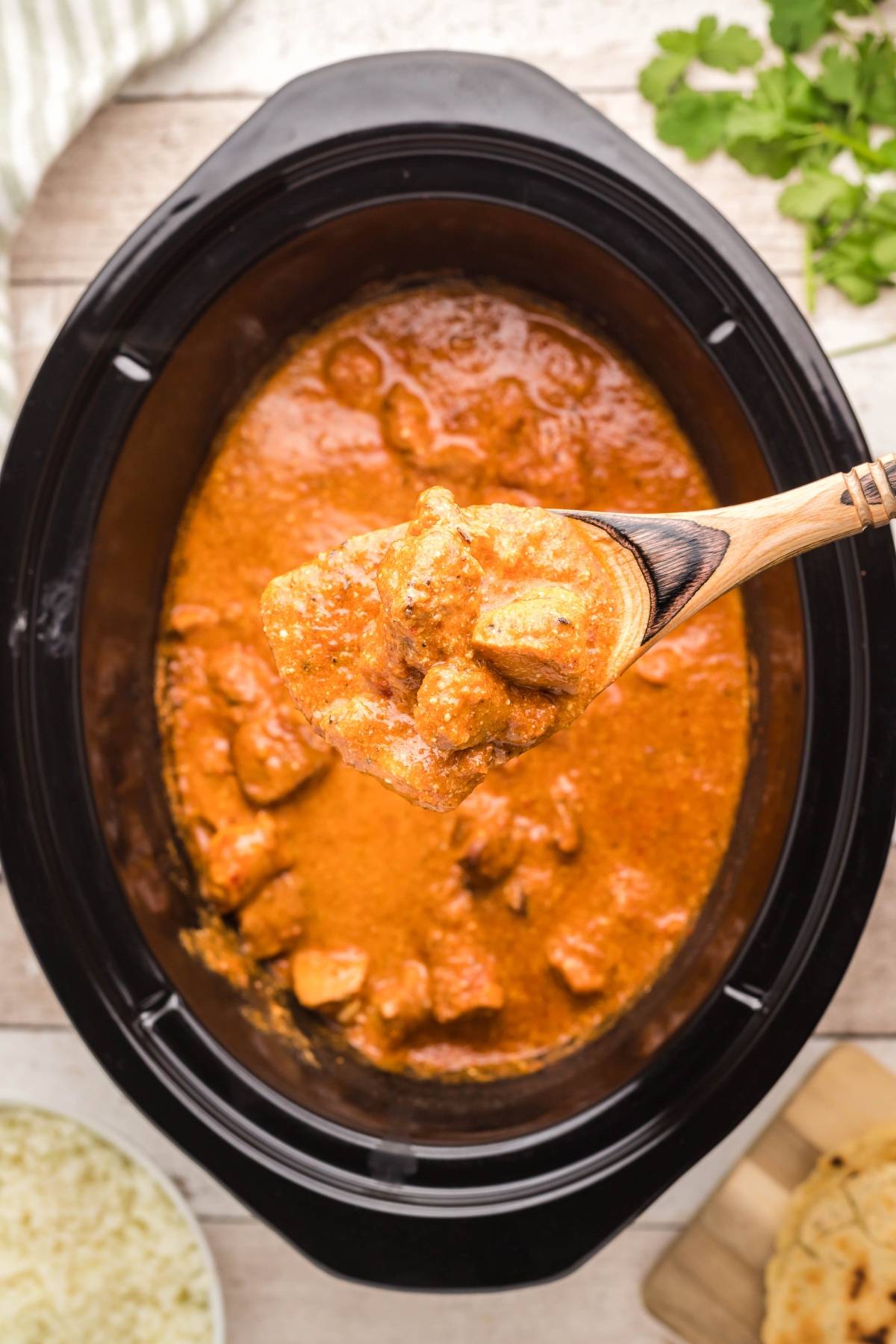 A wooden spoon lifts slow cooker butter chicken tikka masala from a black slow cooker, surrounded by naan and fresh herbs.
