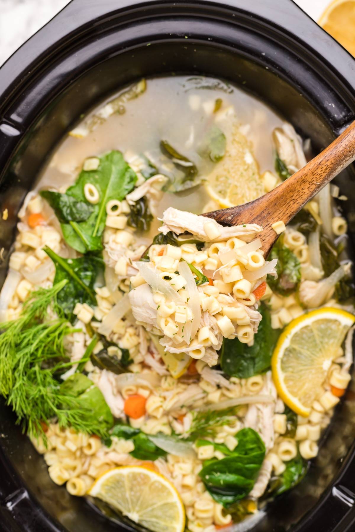 Authentic Greek avgolemono soup with chicken, pasta, spinach, dill, and lemon slices simmered in a slow cooker and stirred with a wooden spoon.