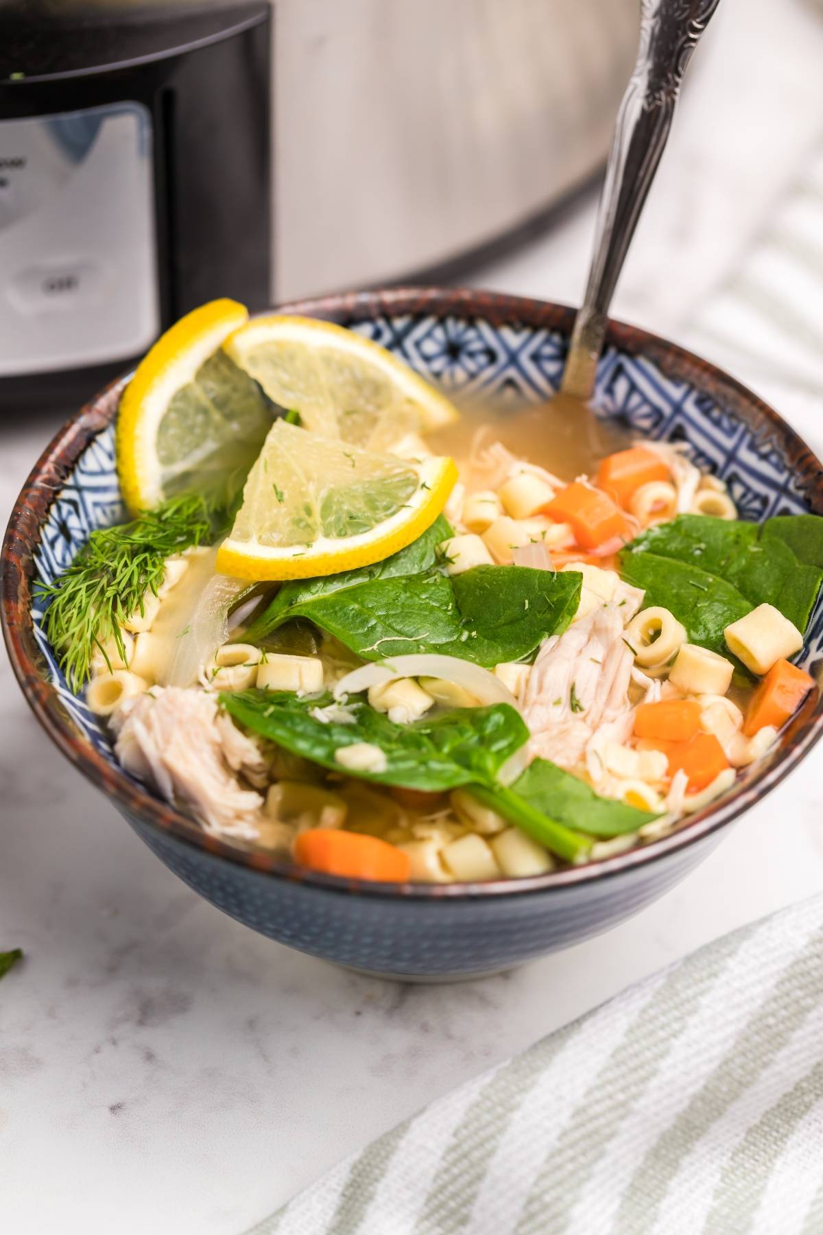 A bowl of authentic Greek soup with chicken, noodles, spinach, carrots, lemon slices, and fresh dill sits next to a slow cooker—perfect for enjoying classic Greek lemon chicken soup at home.