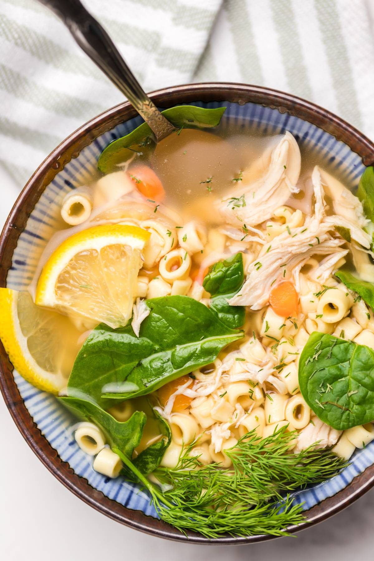 Bowl of authentic Greek soup—chicken noodle avgolemono with spinach, dill, lemon slices, and a spoon on a striped towel background.