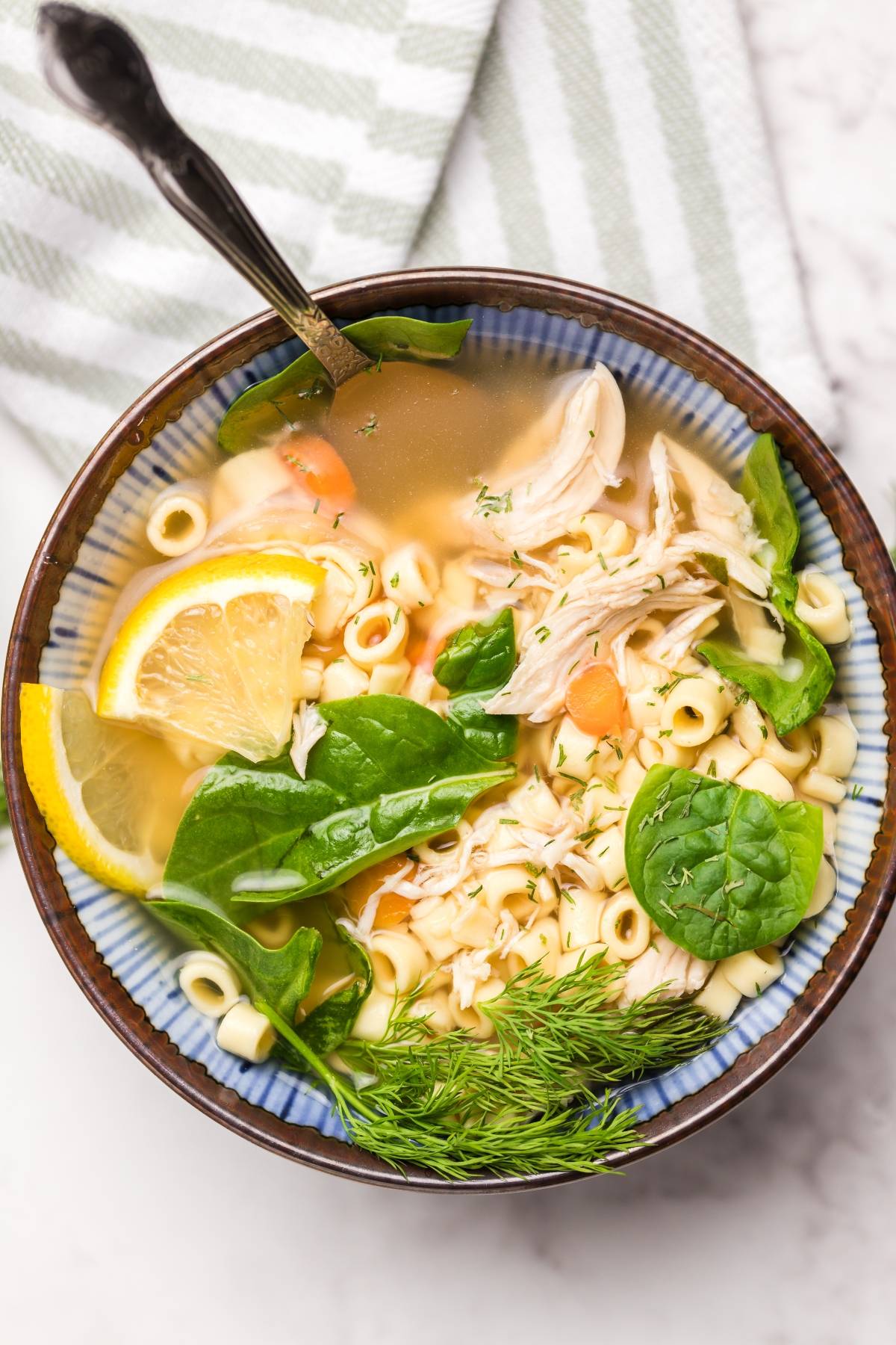Bowl of authentic Greek avgolemono soup with chicken, noodles, fresh spinach, lemon slices, dill, and a spoon, on a striped cloth.