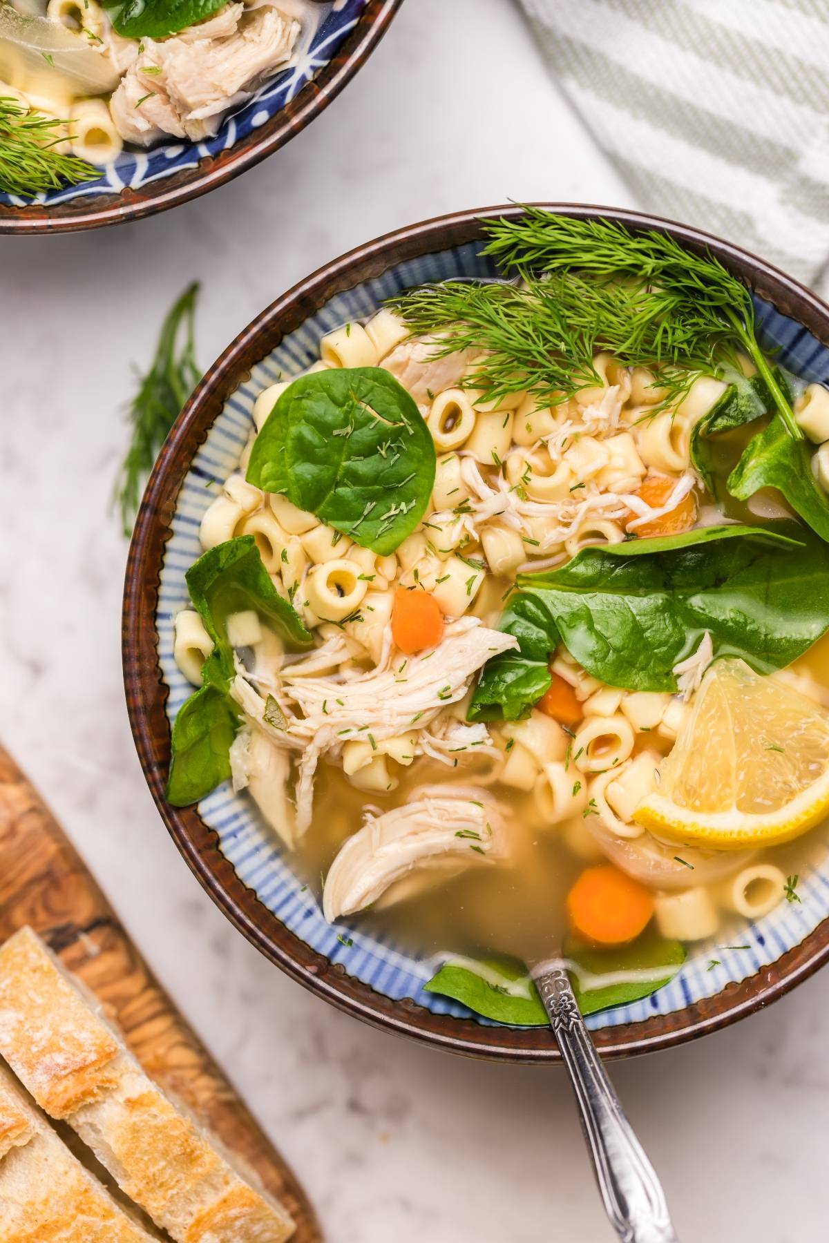 Bowl of Greek lemon chicken soup with carrots, spinach, dill, and a lemon wedge, served with bread on the side—a comforting twist on traditional avgolemono soup.