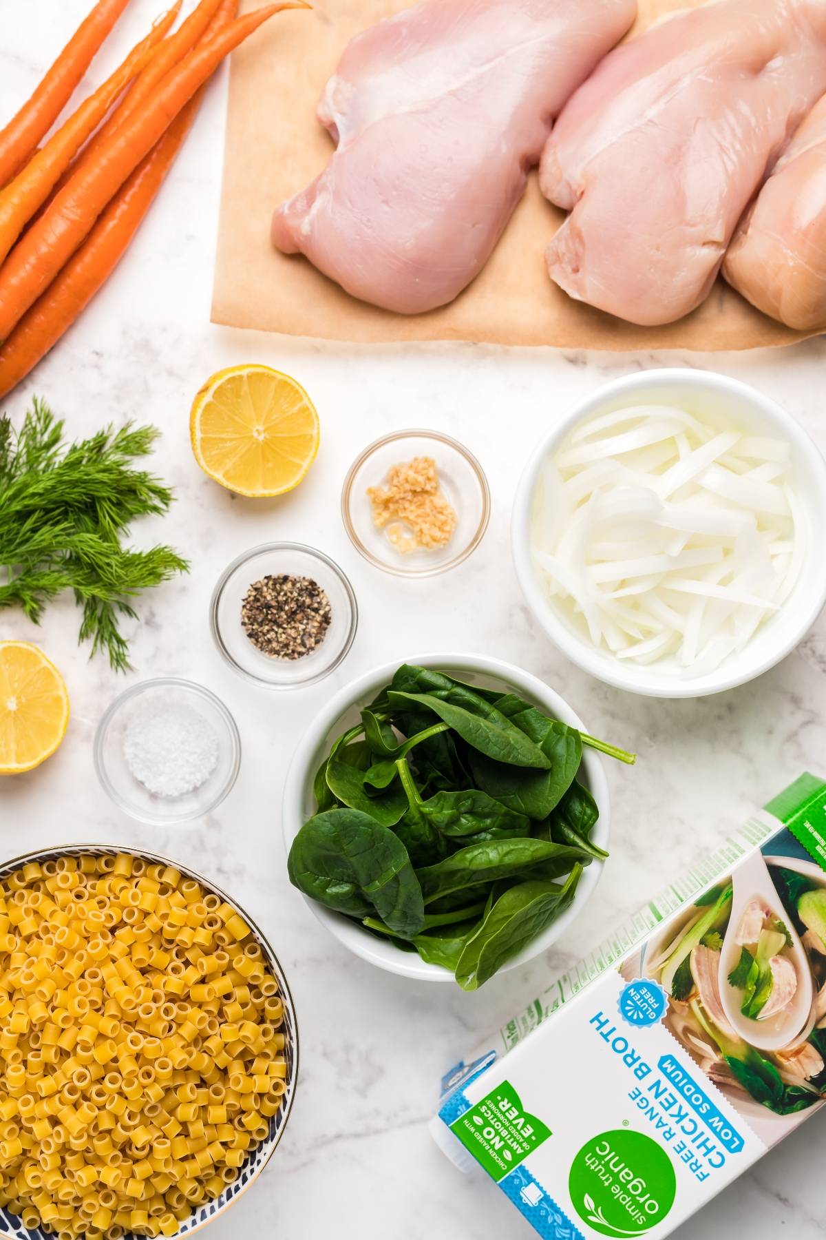 Raw chicken breasts, carrots, lemon, onion, garlic, spinach, pasta, broth, dill, salt, and pepper—everything you need for a hearty slow cooker soup or a classic recipe for chicken avgolemono soup.