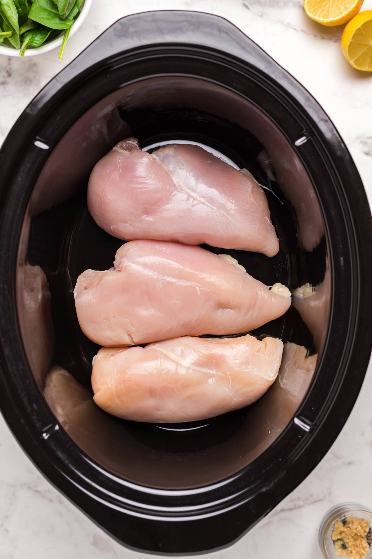Four raw chicken breasts placed inside a black slow cooker, with lemon and spinach visible nearby—perfect for starting a recipe for chicken avgolemono soup, the classic Greek lemon chicken soup.