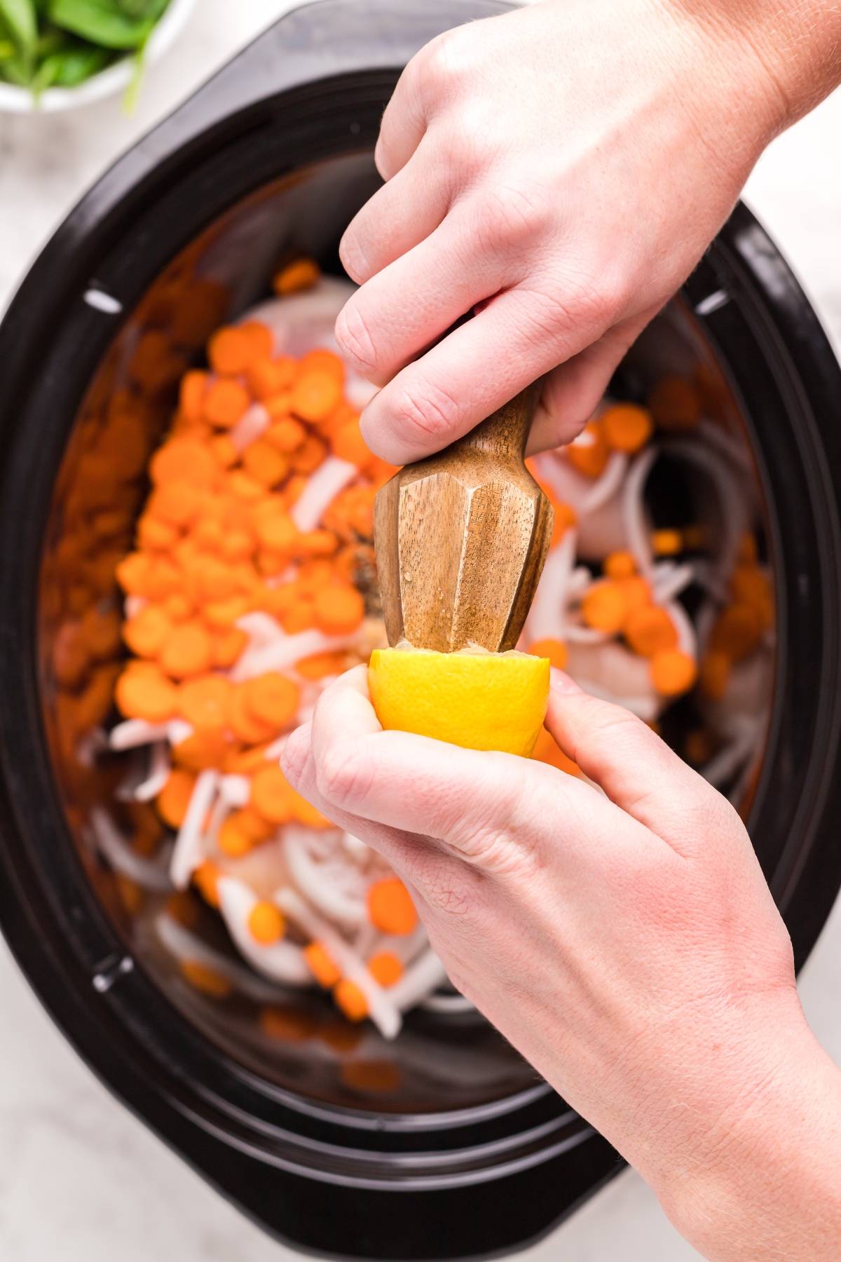 Hands squeezing a lemon over sliced carrots and onions in a slow cooker, preparing the base for an authentic Greek soup.