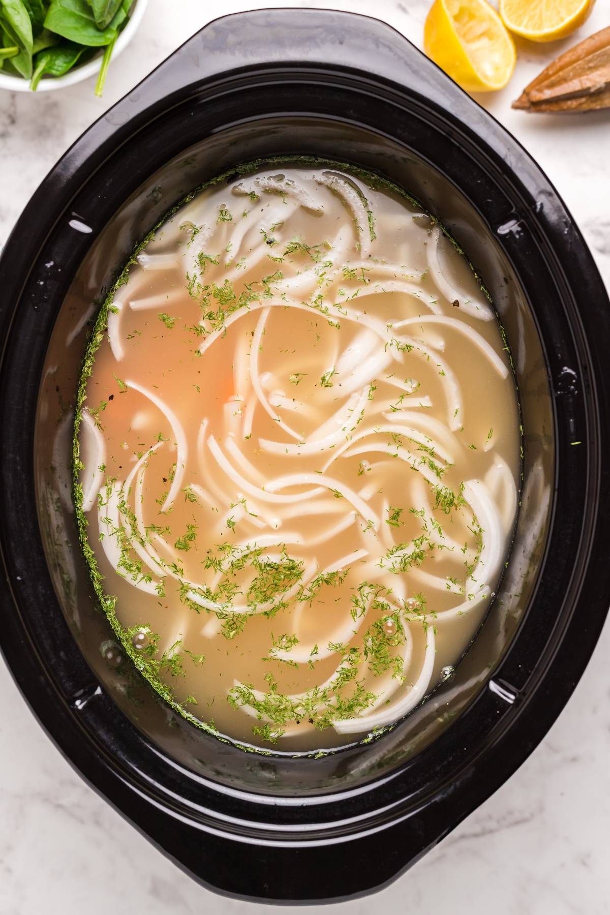 A slow cooker filled with broth, sliced onions, and chopped herbs on a marble countertop—perfect for preparing an authentic Greek soup like avgolemono soup.
