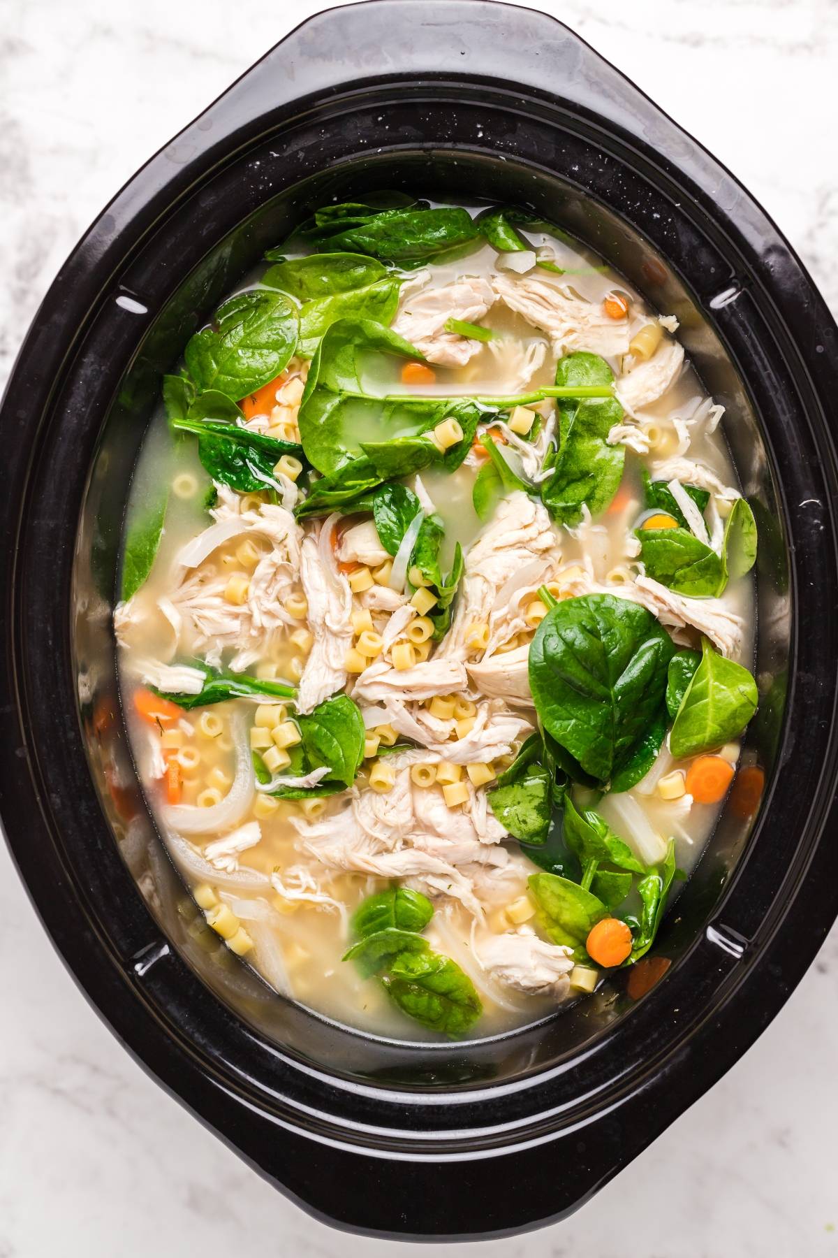 Shredded chicken soup with spinach, carrots, and pasta in a black slow cooker on a light surface—an easy slow cooker soup inspired by authentic Greek avgolemono flavors.