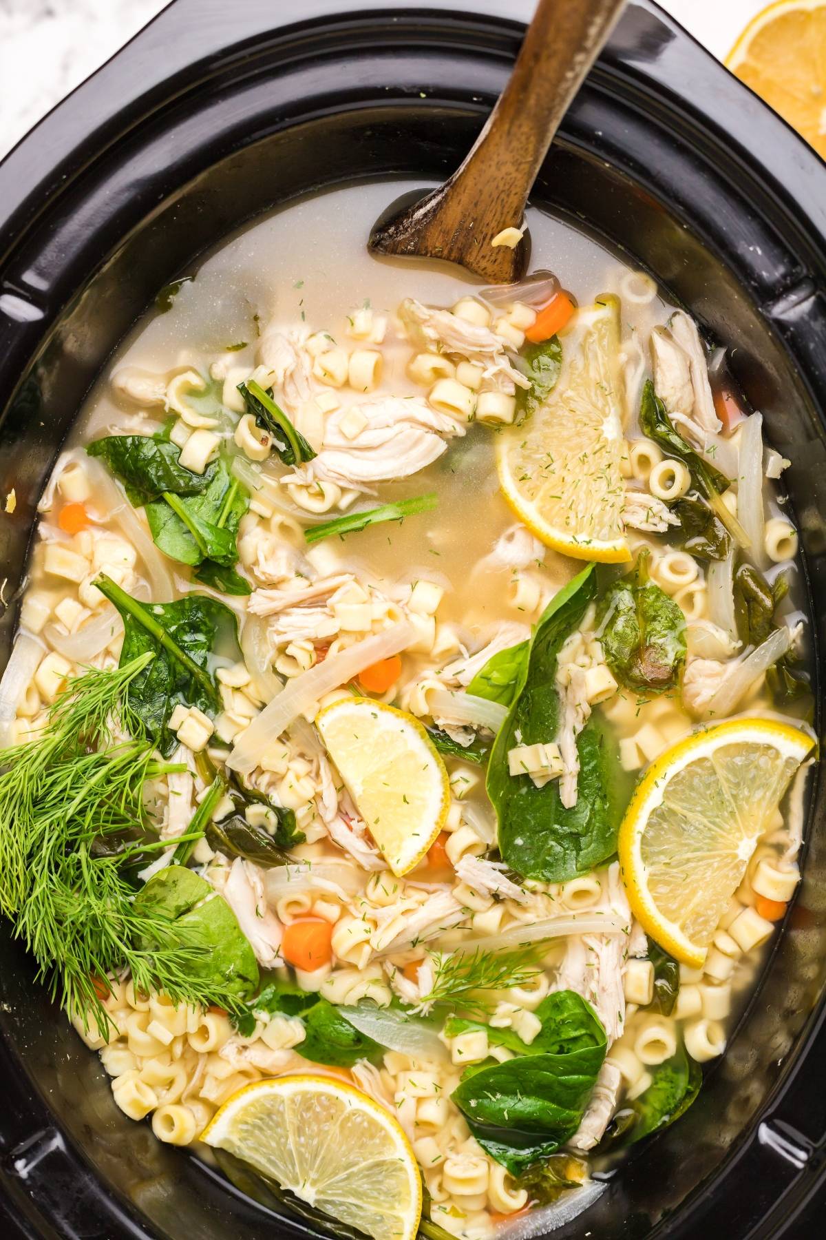 Chicken noodle soup in a slow cooker with spinach, carrots, lemon slices, and a wooden spoon—a cozy twist on Greek lemon chicken soup for authentic Greek soup lovers.