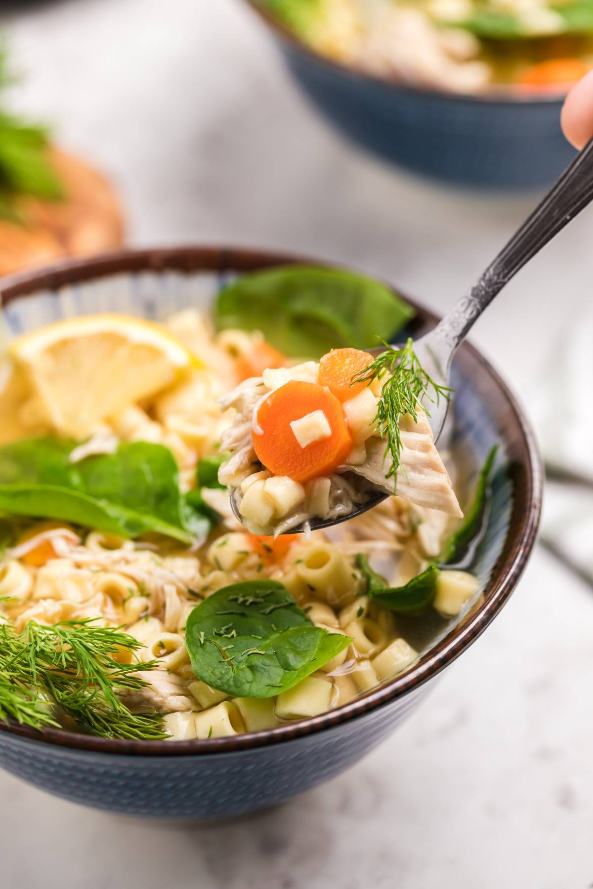 A spoonful of Greek lemon chicken soup with carrots, spinach, pasta, and dill held above a bowl—an inviting taste of authentic avgolemono soup.