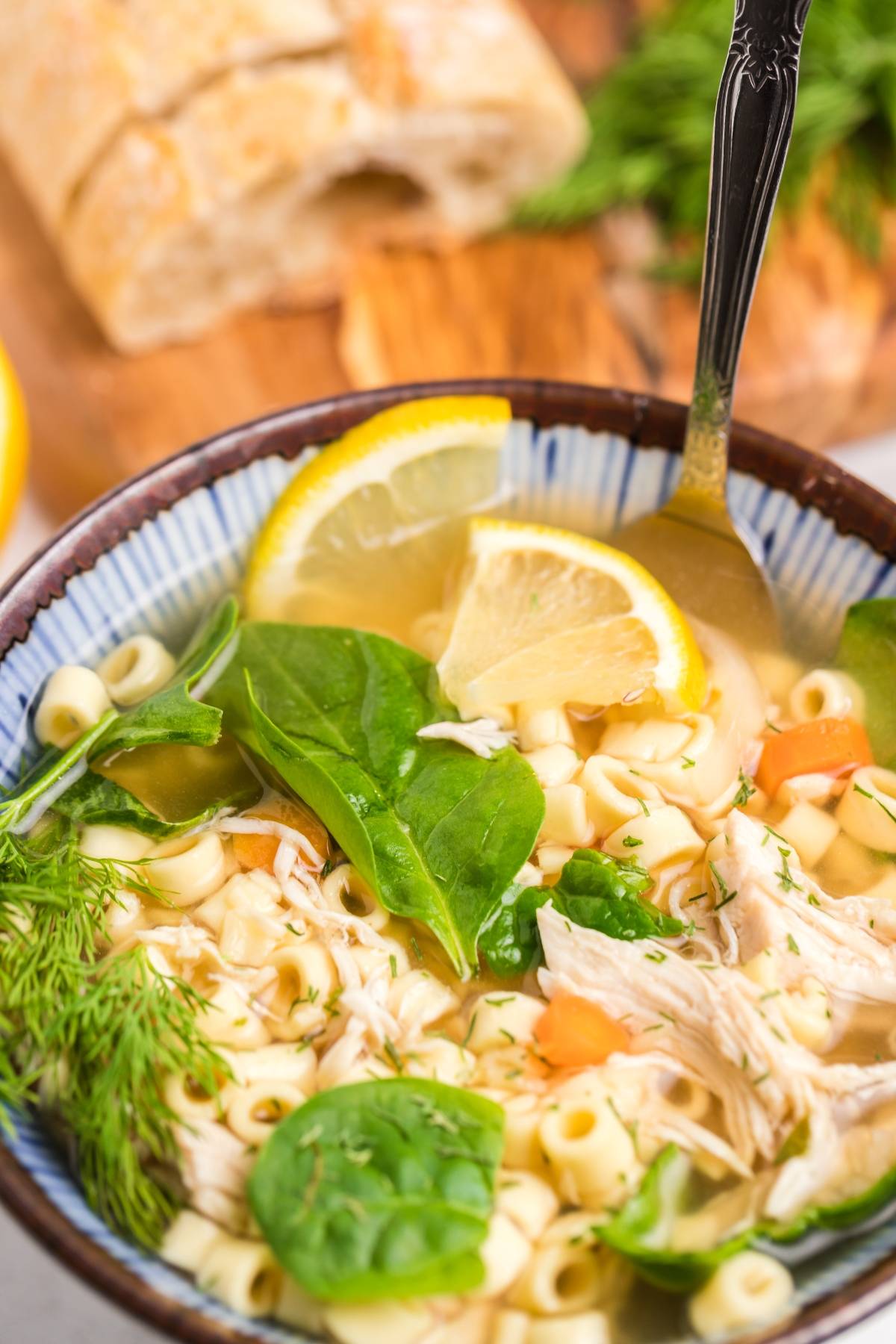 A bowl of Greek lemon chicken soup, also known as avgolemono soup, with spinach and lemon slices, served alongside fresh bread in the background.