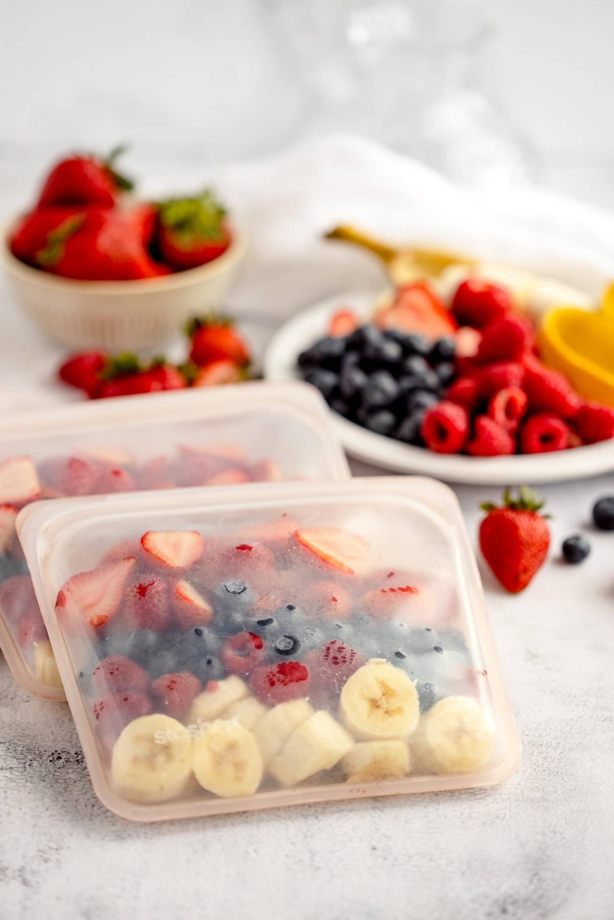Reusable bags filled with sliced bananas, strawberries, and blueberries—perfect for make ahead smoothie mix; fresh fruit in the background.