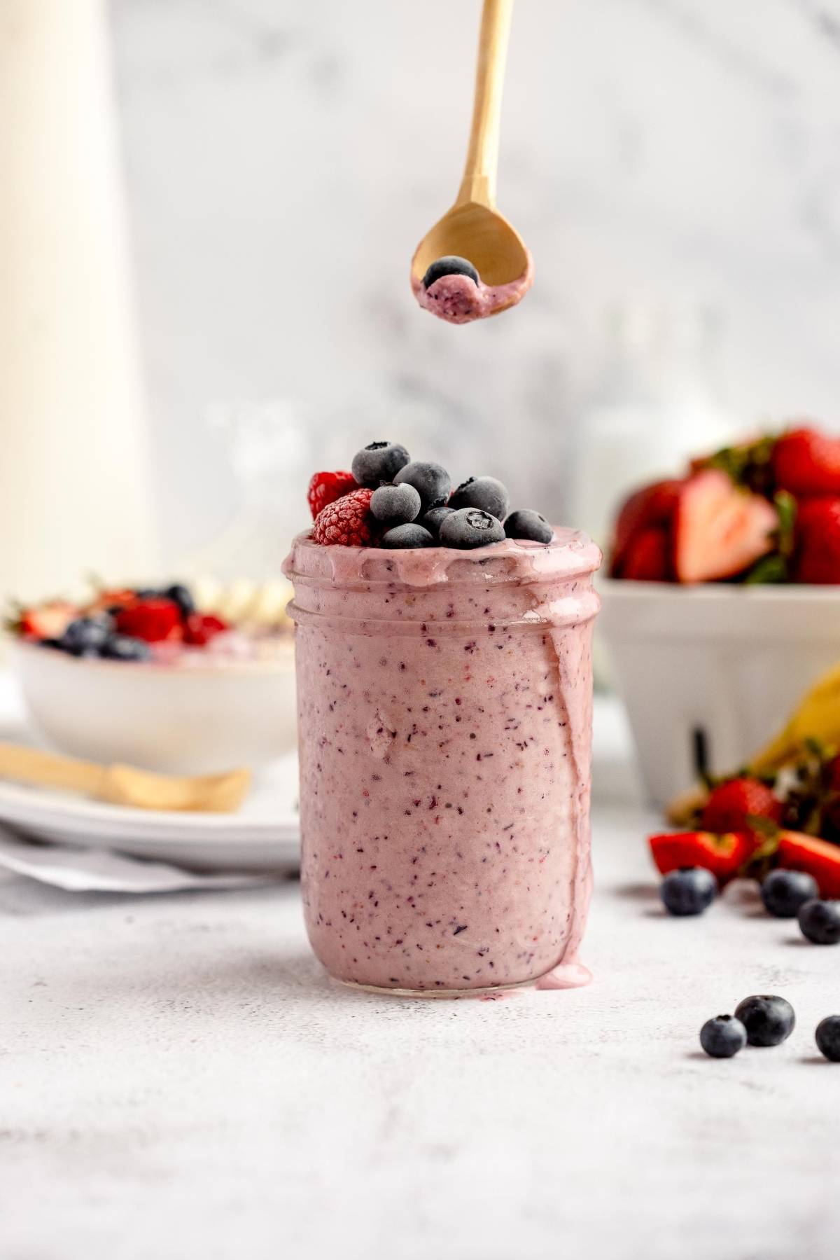 A jar of berry smoothie topped with fresh berries; a spoon with blueberries is held above it—perfect inspiration for a Smoothie Freezer Packs - DIY Meal Prep.
