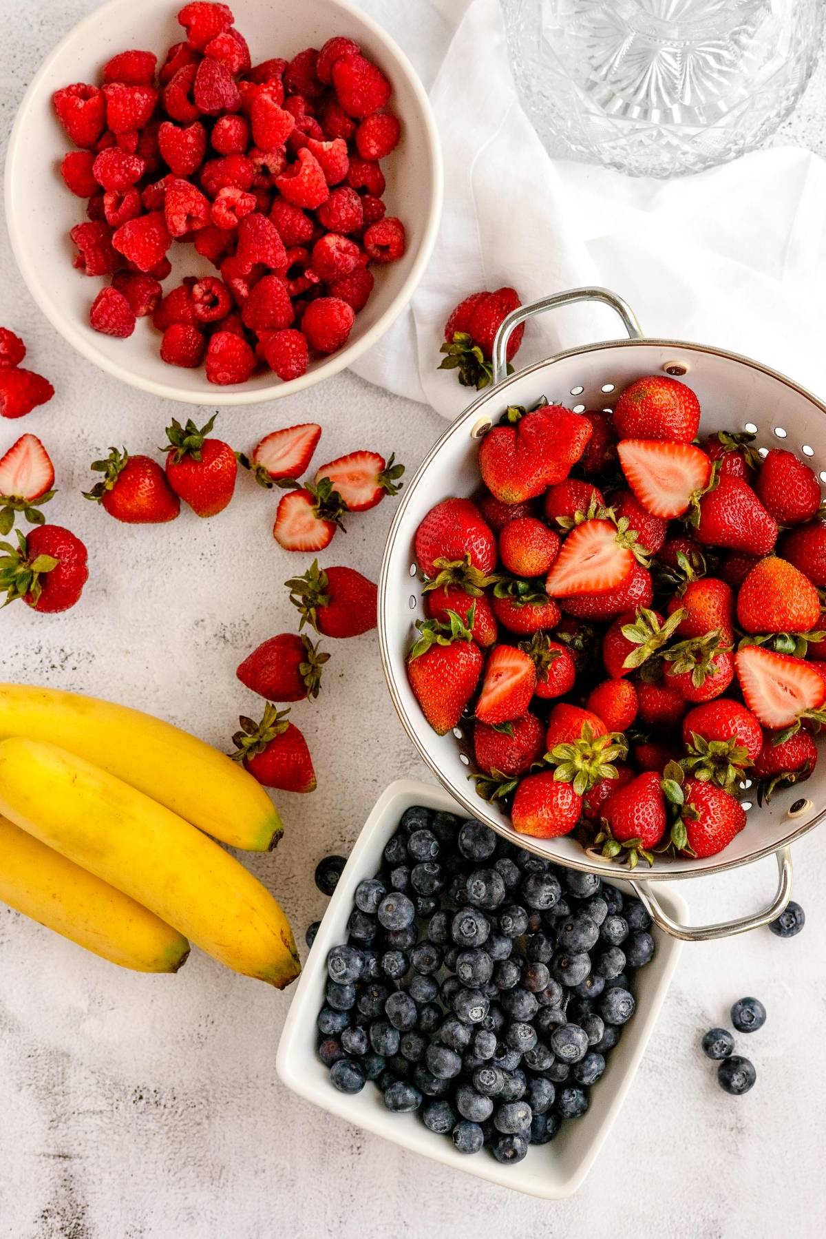 Bowls of raspberries, strawberries, blueberries, and bananas on a white surface—perfect for creating a homemade smoothie pack or DIY Meal Prep for a make ahead smoothie mix.