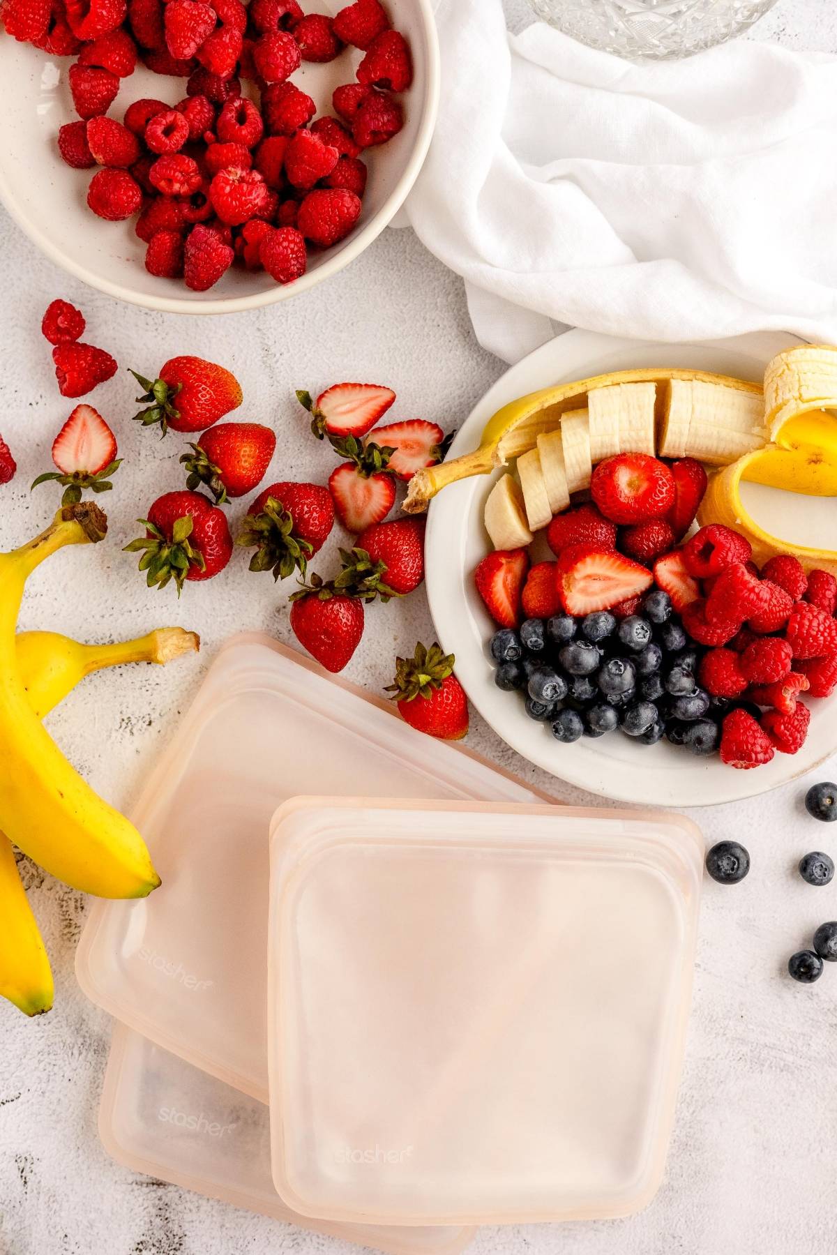 Fresh berries, banana, and strawberries on plates with reusable silicone bags and a white cloth—perfect for creating a homemade smoothie pack or DIY Meal Prep make ahead smoothie mix.