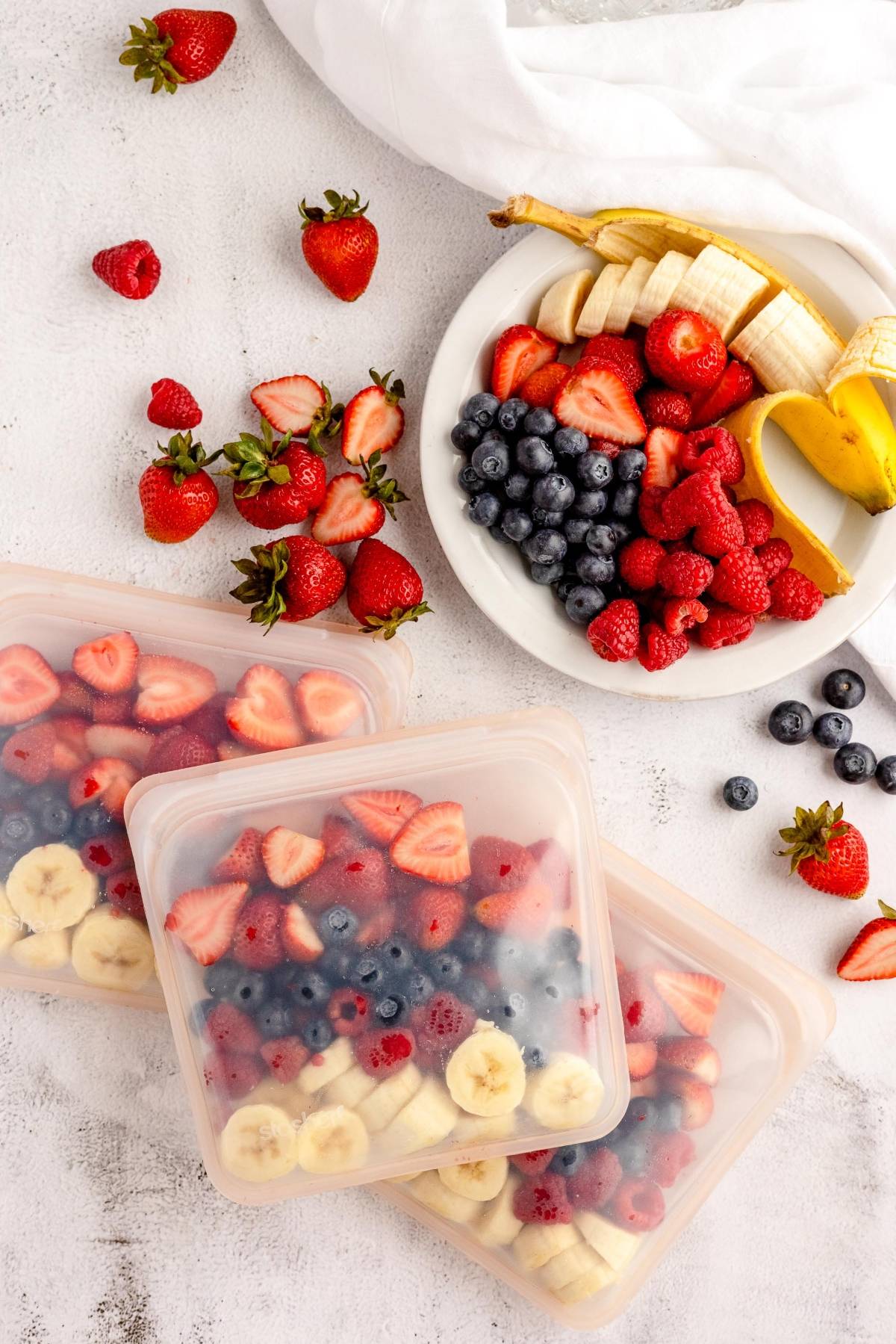 A bowl of mixed berries and banana sits beside three frozen smoothie bags filled with sliced strawberries, bananas, and blueberries—perfect for DIY Meal Prep and quick smoothie freezer packs.