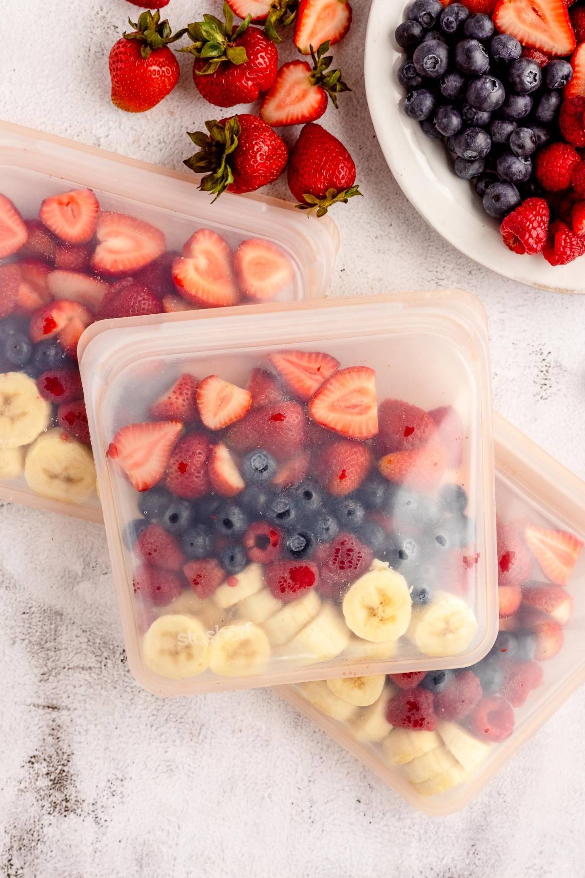 Sliced strawberries, bananas, and blueberries in reusable bags for a make ahead smoothie mix, with fresh berries on a plate and table—an easy DIY meal prep solution.